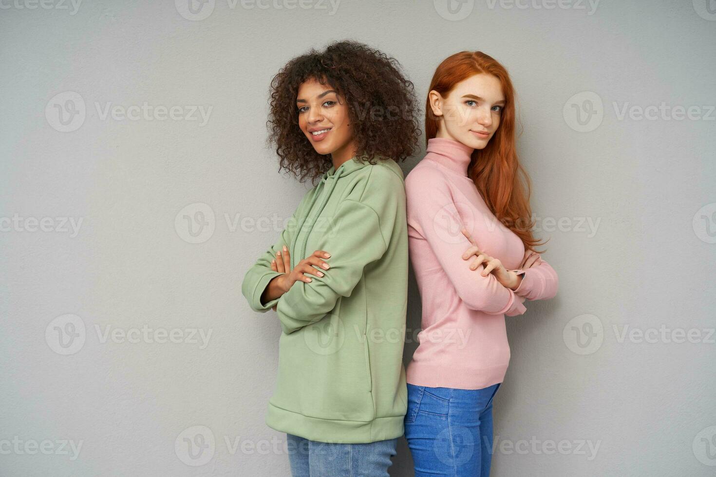 Indoor shot of beautiful young positive women keeping their hands crossed while standing back to back over grey background, looking at camera with pleasant smile photo