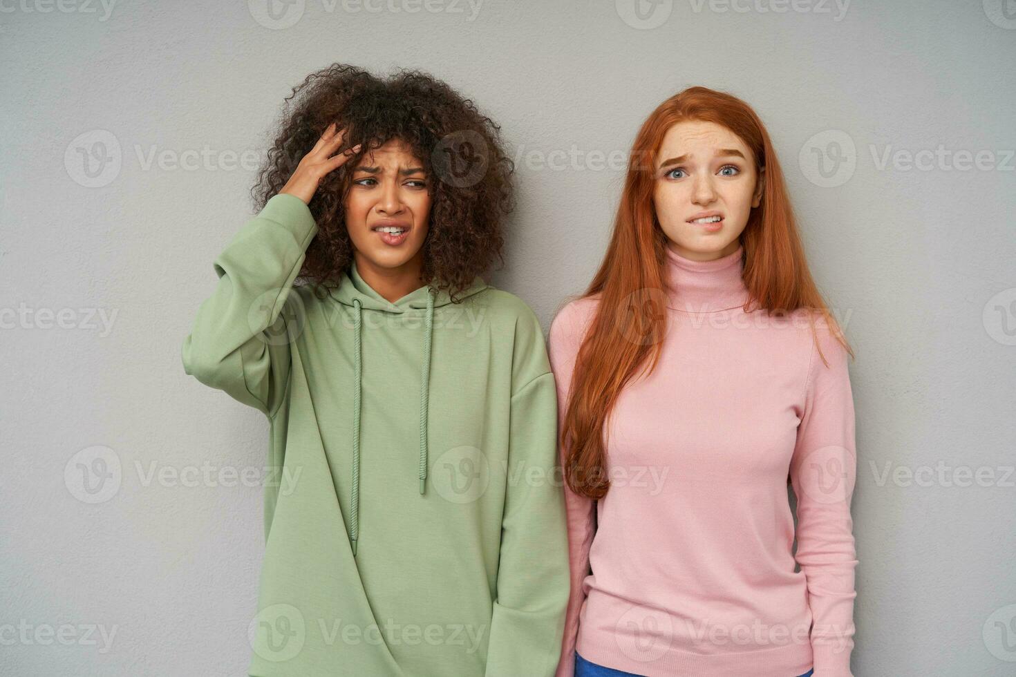 Studio photo of bemused young pretty ladies biting worringly their underlips and frowning confusedly eyebrows while standing over grey background in casual clothes