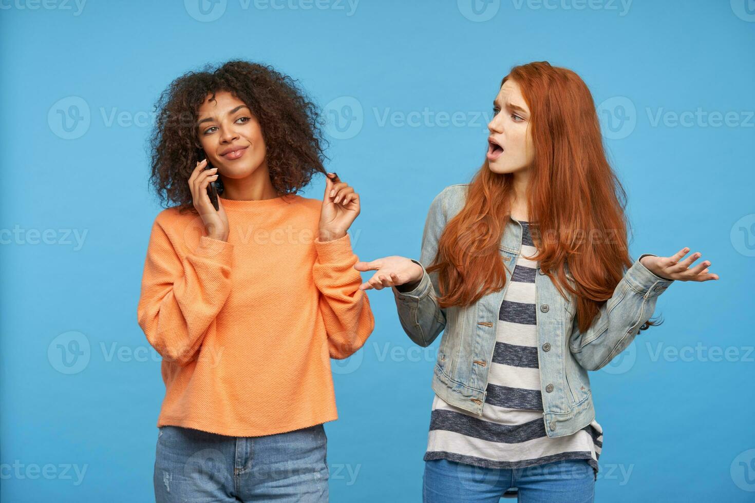 Studio photo of bewildered young long haired redhead female raising confusedly palms while looking at her cheerful dark haired curly dark skinned lady with smartphone, isolated over blue background