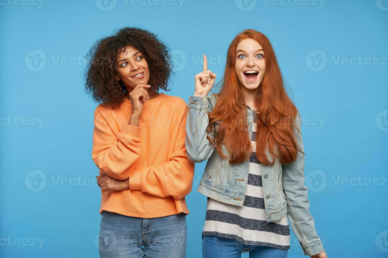 Overjoyed young lovely long haired redhead woman raising forefinger while looking excitedly atcamera, posing over blue background with thoughtful positive curly dark skinned brunette lady photo