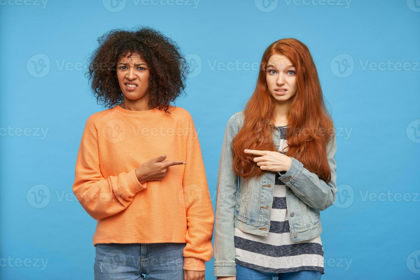 Studio photo of displeased young pretty women keeping their forefingers raised while looking at camera with pout, posing over blue background in casual clothes