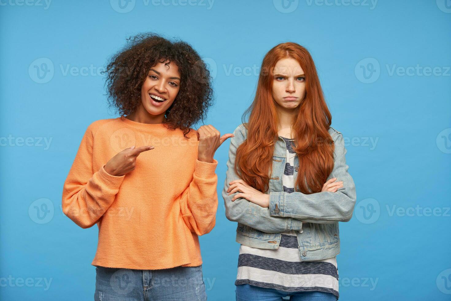 Joyful young lovely curly dark skinned brunette female in orange knitted sweater showing with raised hands on her offended redhead girlfriend, isolated over blue background photo