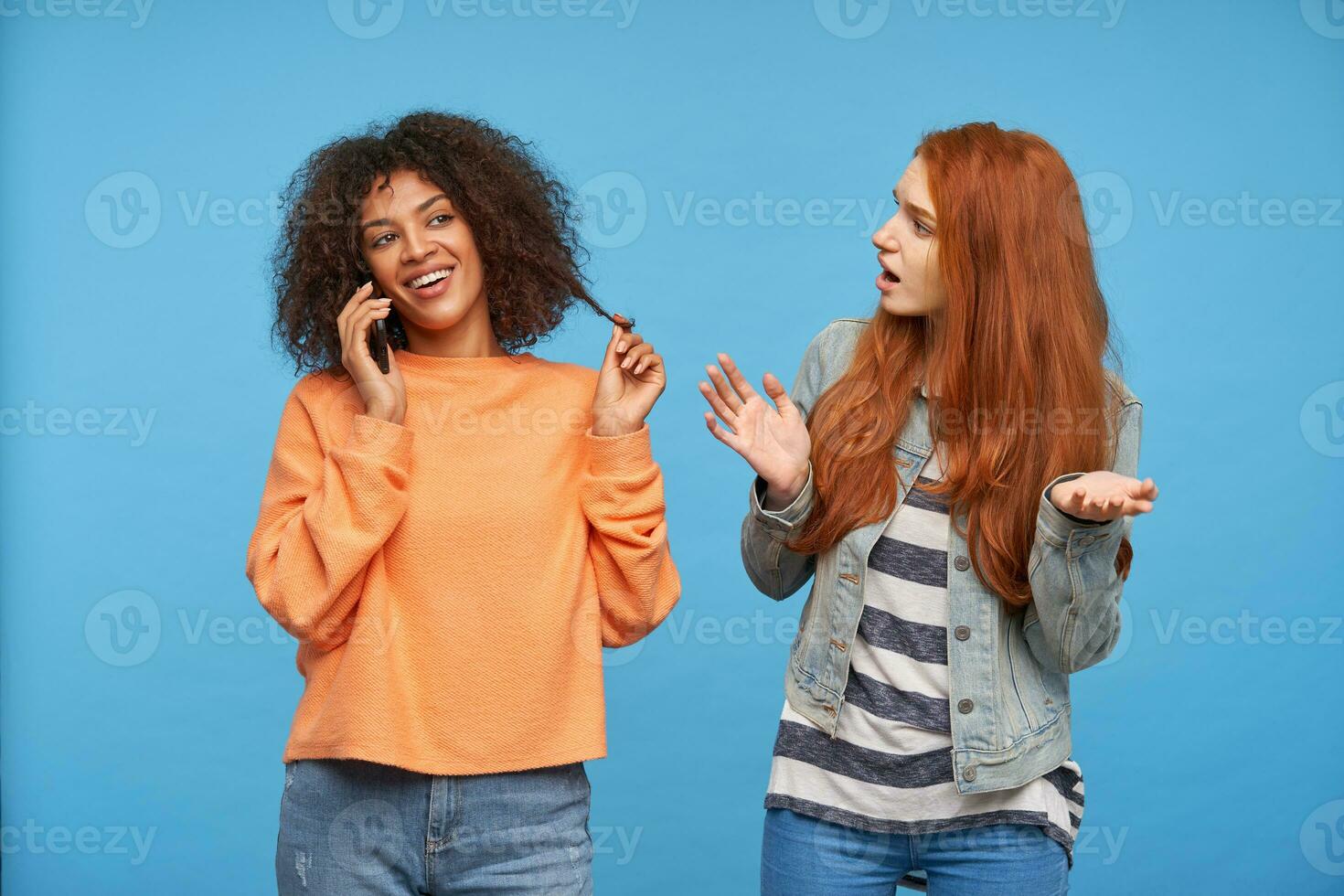 alegre joven bonito Rizado oscuro desollado morena hembra sonriente felizmente mientras teniendo agradable teléfono conversación, posando terminado azul antecedentes con confuso pelirrojo atractivo mujer foto