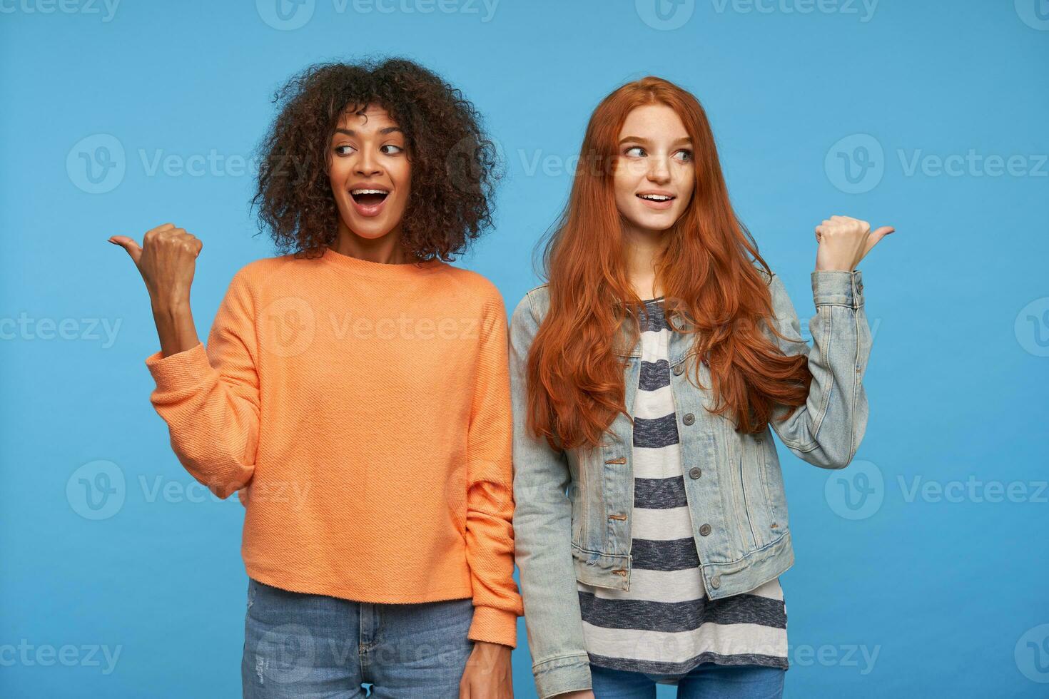 Indoor photo of attractive agitated women pointing in different sides with raised thums and excited faces, standing against blue background in casual clothes