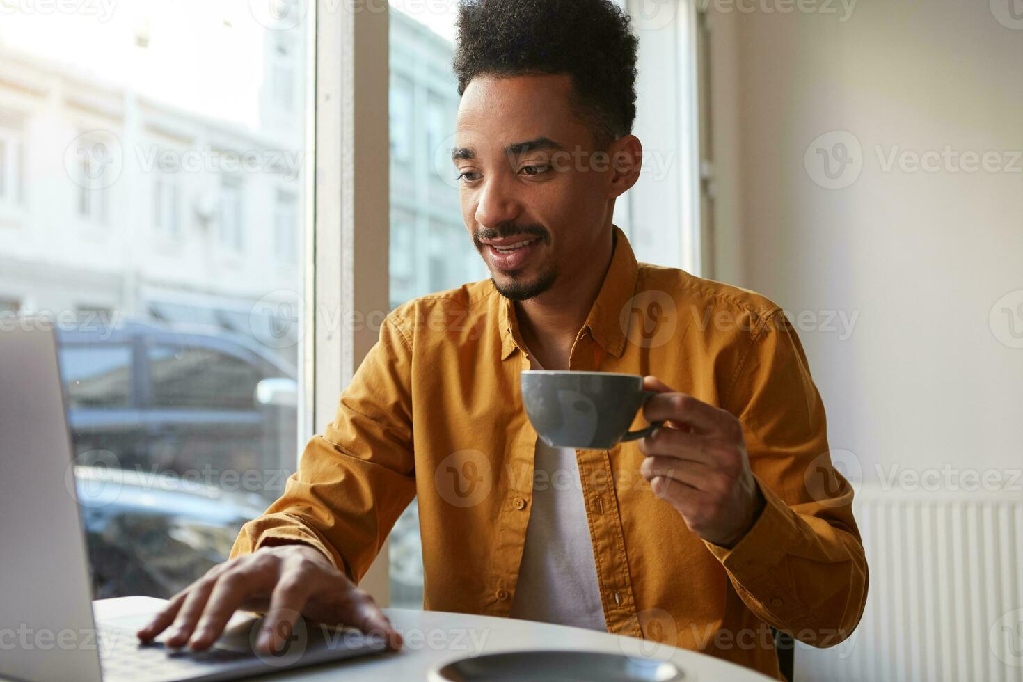 African American boywears in yellow shirt, sitting in a cafe and working on a laptop, drinks coffee, looking for a route for a new journey, enjoys the day. photo