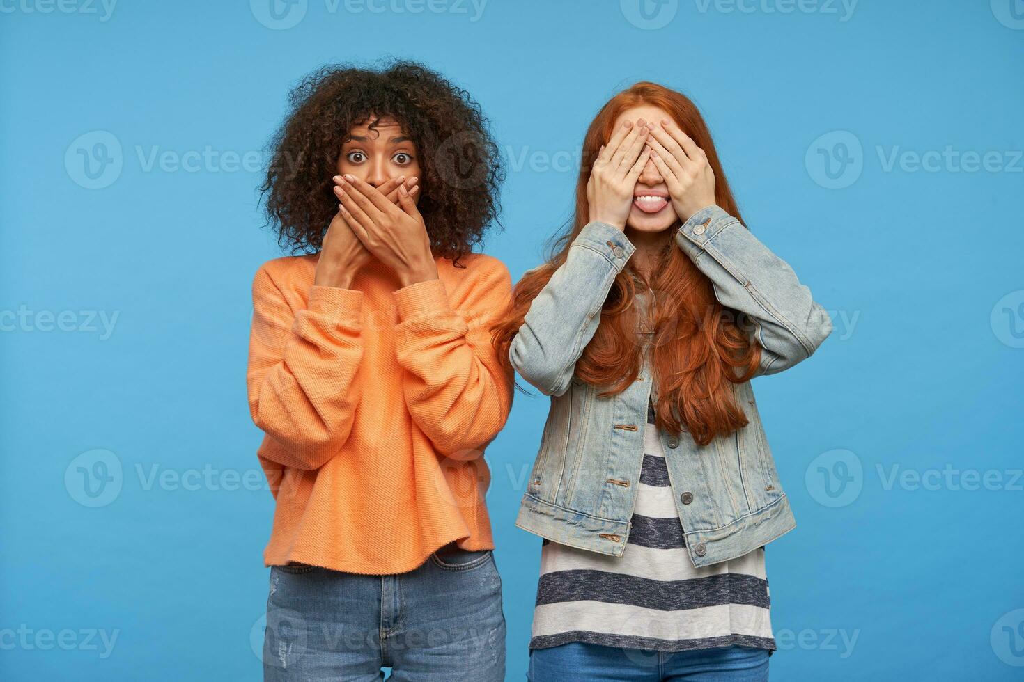 Joyful lovely young redhead lady covering her eyes and showing happily tongue while posing over blue background with amazed young dark haired curly dark skinned woman photo