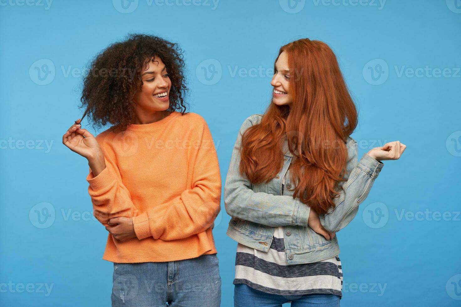 Happy attractive ladies dressed in casual clothes keeping their hands raised while looking cheerfully on each other with wide charming smiles, isolated over blue background photo