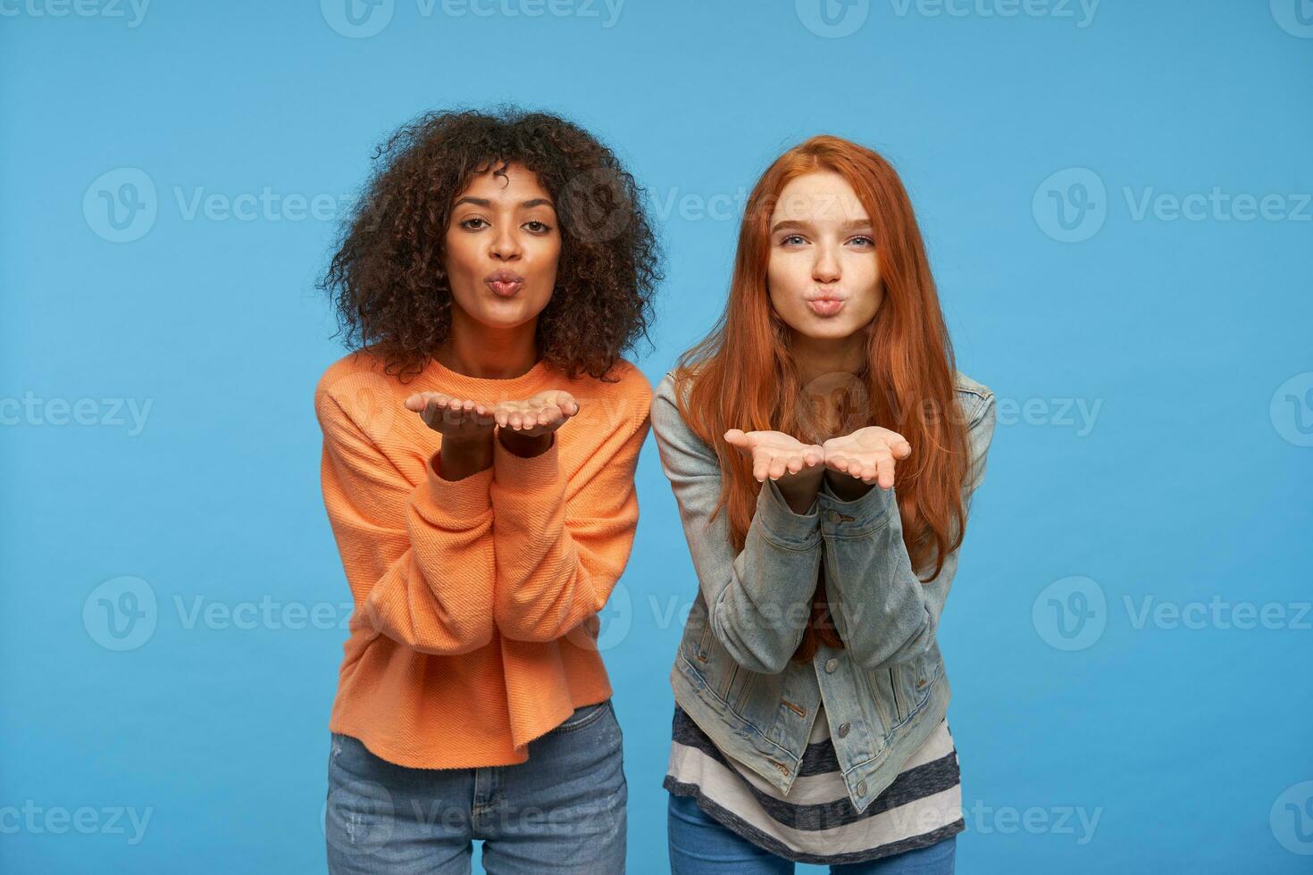 Portrait of charming positive pretty young women raising their palms while posing over blue background, folding their lips and blowing air kiss to camera photo