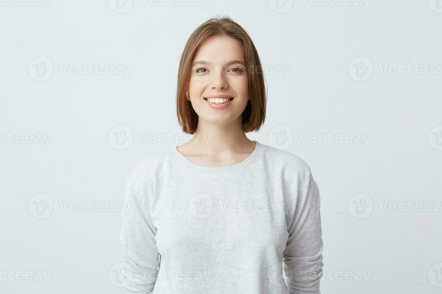 beautiful woman expresses happy emotions, has broad pleasant smile, wears white long-sleeve and feels satisfied. Isolated over white background photo