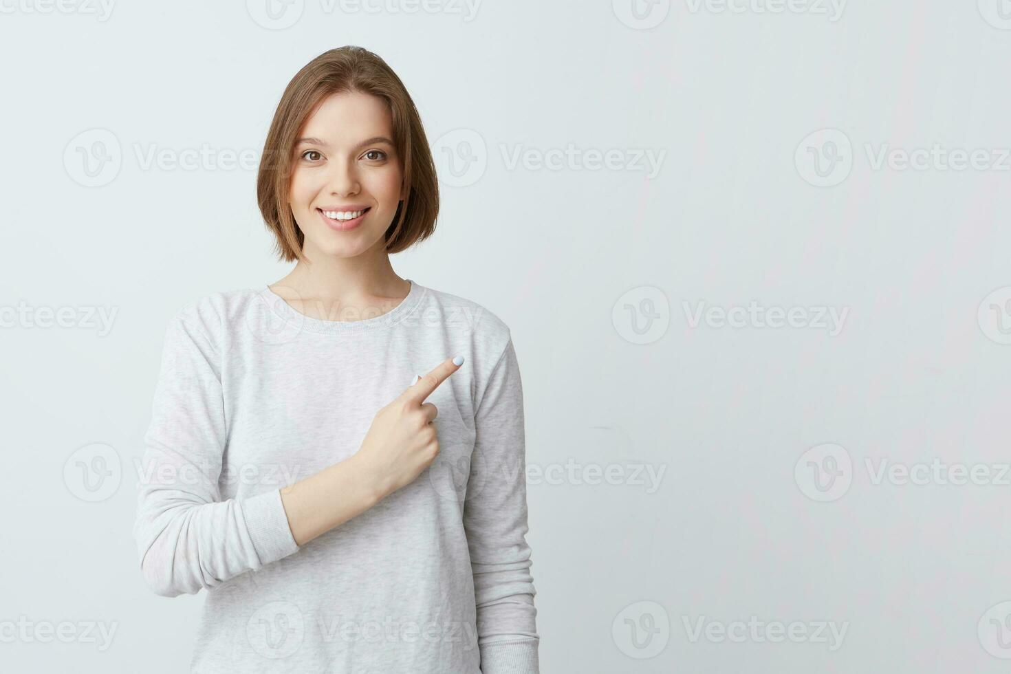sonriente atractivo joven mujer en manga larga en pie y señalando a el lado aislado terminado blanco antecedentes siente contento y confidente foto