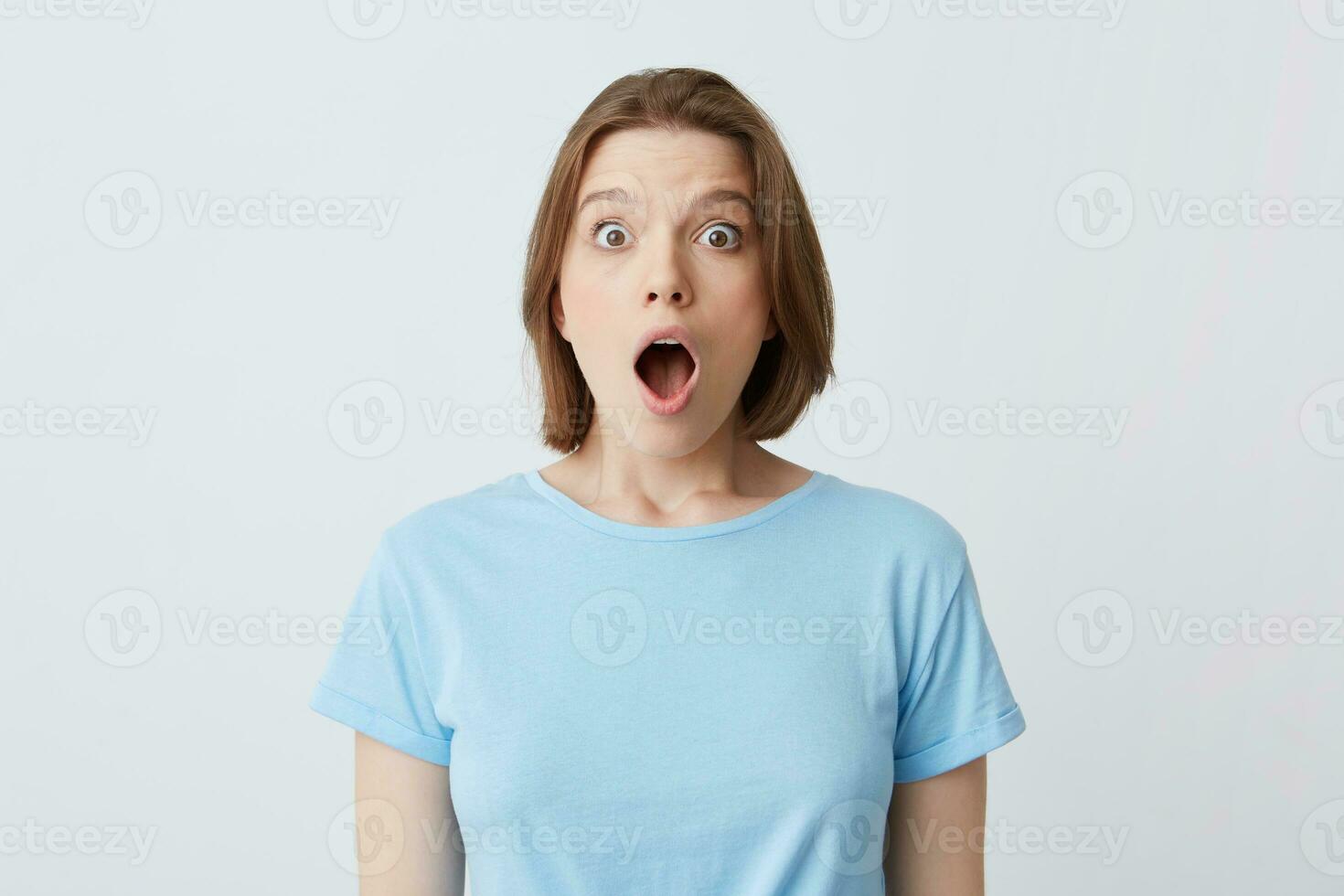 Closeup of shocked stunned young woman in blue t shirt with opened mouth feels amazed and looks directly in camera isolated over white background photo
