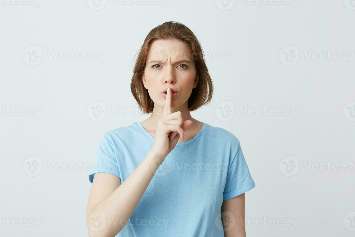 Closeup of serious unhappy young woman in blue t shirt frowning face and showing silence sign isolated over white background Feels irritated and angry photo