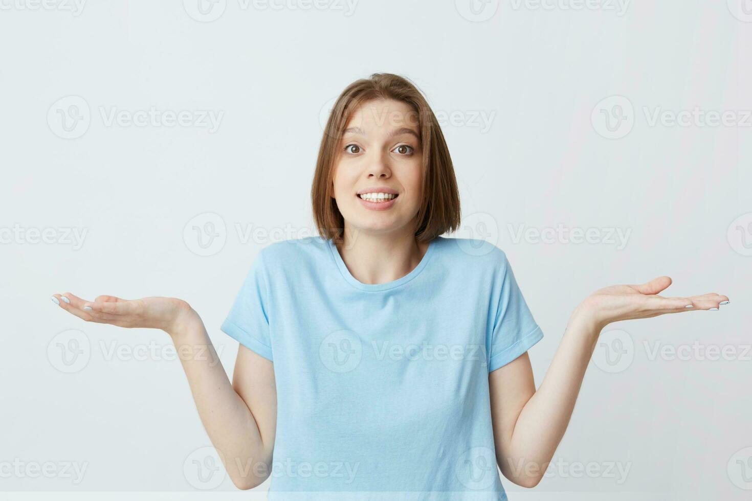 Portrait of confused cute young woman in blue t shirt shrug her shoulders and hold copy space on both palms isolated over white background photo