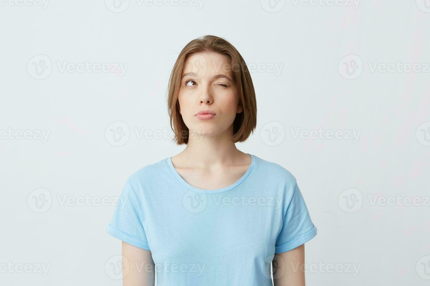 Thoughtful concentrated young woman in blue t shirt with one closed eyes thinking and trying to remember answers for the questions on exam isolated over white wall Looking to the side and up photo