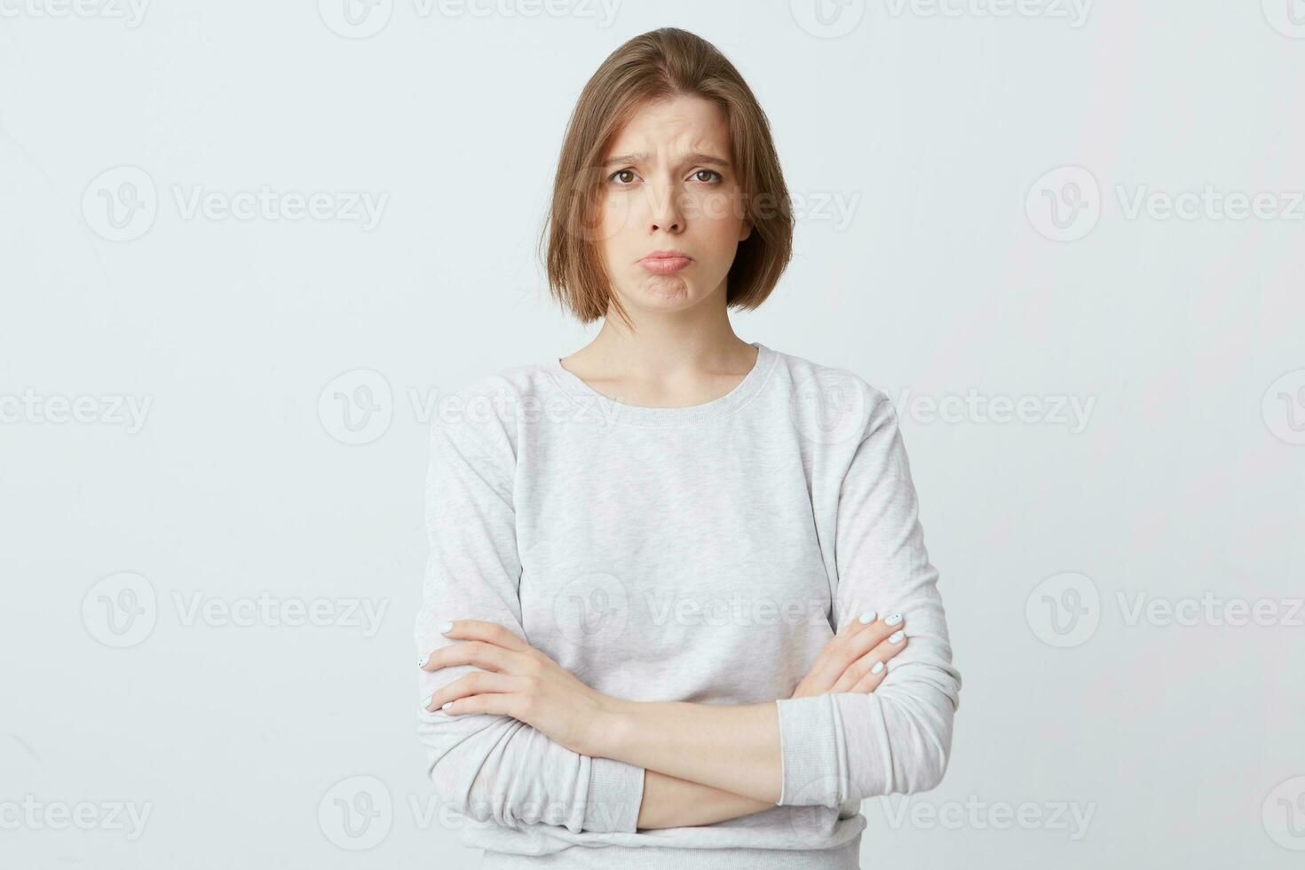 Closeup of sad unhappy pretty young woman in long-sleeve stands with hands folded and feels displeased isolated over white background Looks directly in camera photo