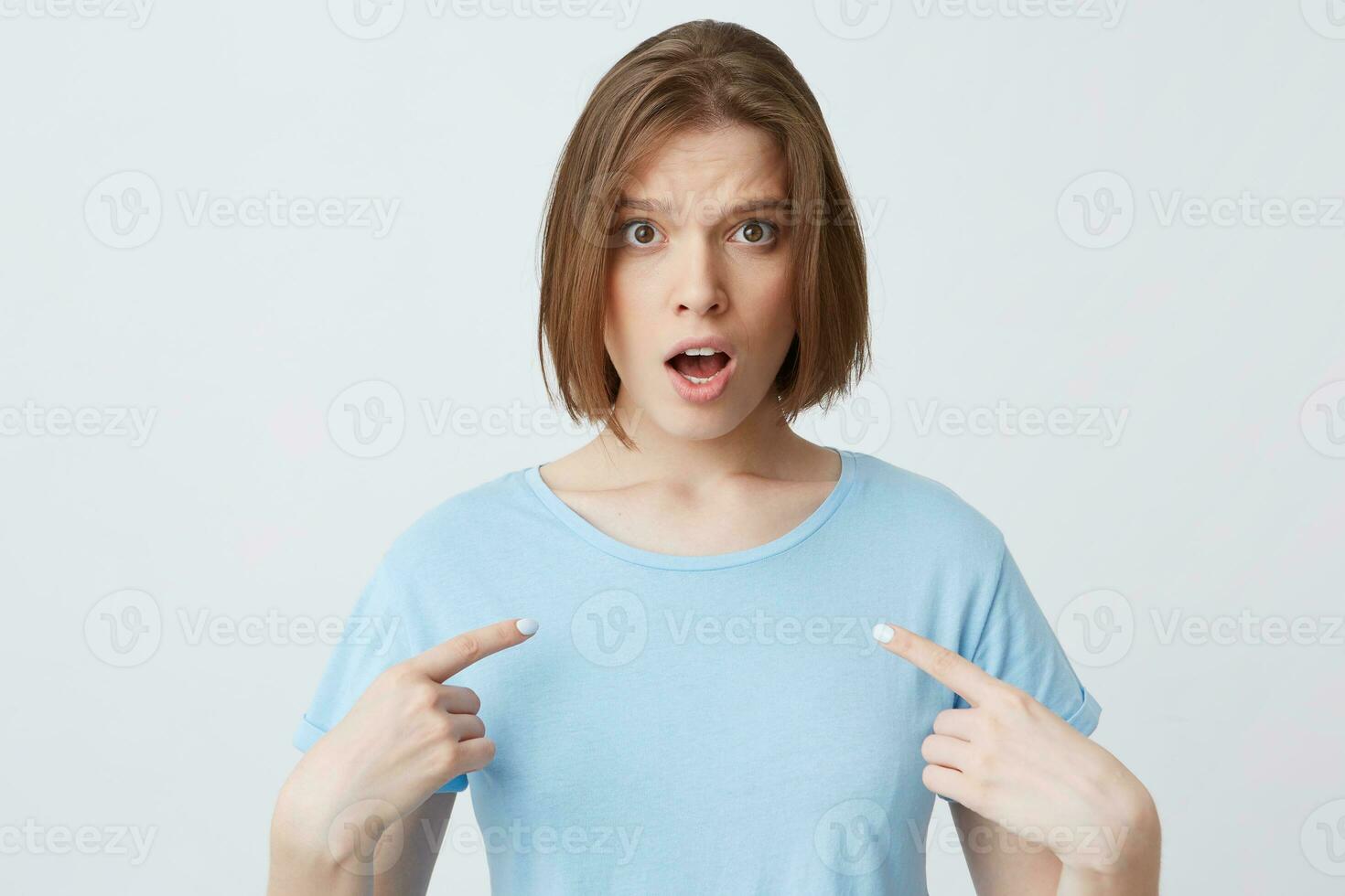 Amazed shocked young woman in blue t shirt with opened mouth looks stunned and points at herself with fingers on both hands isolated over white background photo