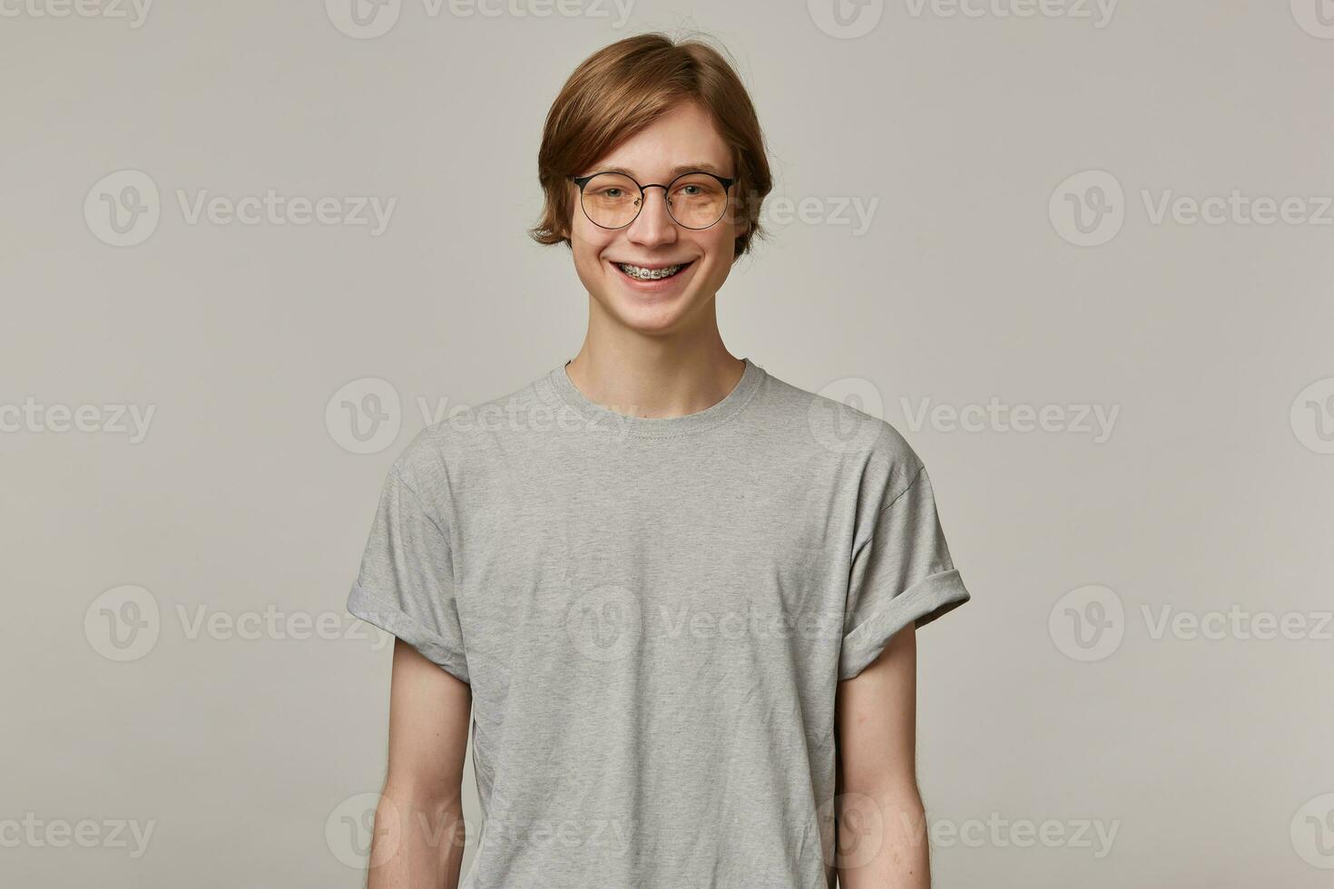 Portrait of cheerful, adult male with blond hair. Wearing grey t-shirt, glasses and has braces. People and emotion concept. Watching at the camera and smiling isolated over grey background photo