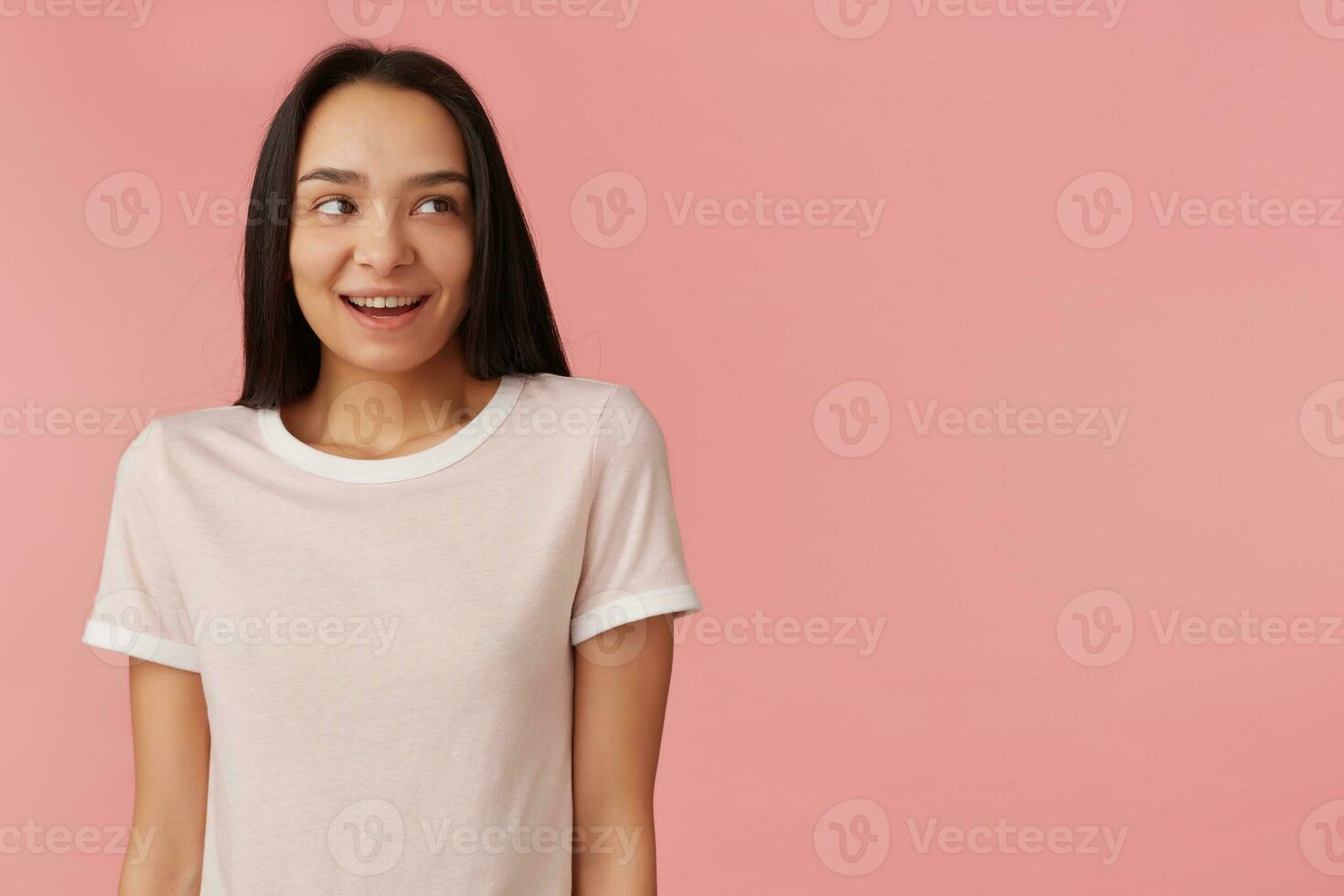 retrato de atractivo, adulto niña con negro largo cabello. vistiendo blanco camiseta. encogidamente sonriente y se encoge de hombros espalda. acecho a el Derecha a Copiar espacio, aislado terminado pastel rosado antecedentes foto