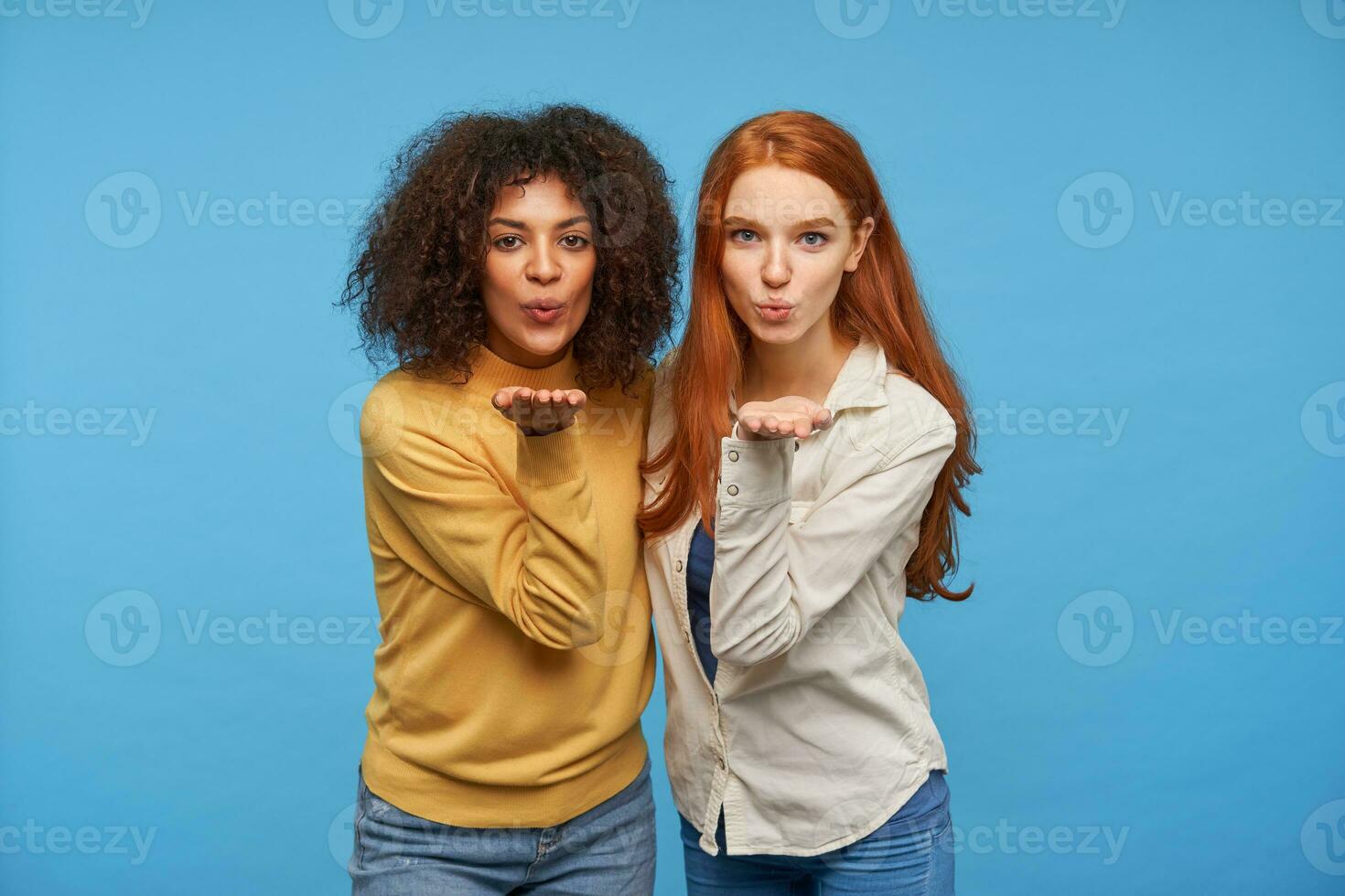 Couple of charming positive young women dressed in casual wear raising their palms up and pursing lips while blowing air kiss at camera, isolated over blue background photo