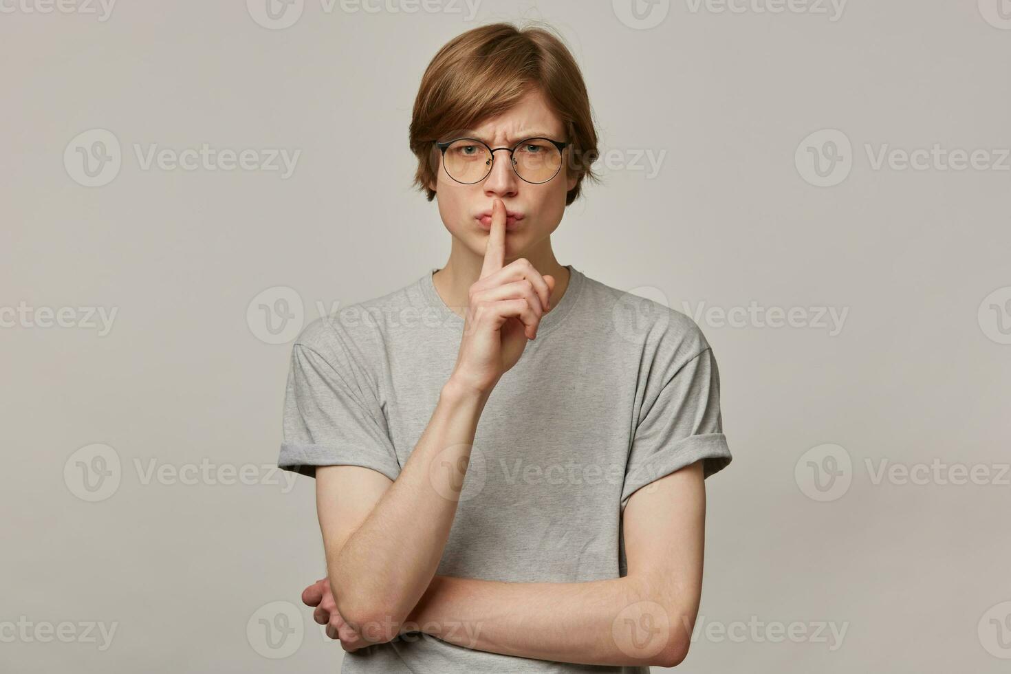 Portrait of handsome, angry male with blond hair. Wearing grey t-shirt and glasses. People and emotion concept. Showing silence sign and frowns. Watching at the camera isolated over grey background photo