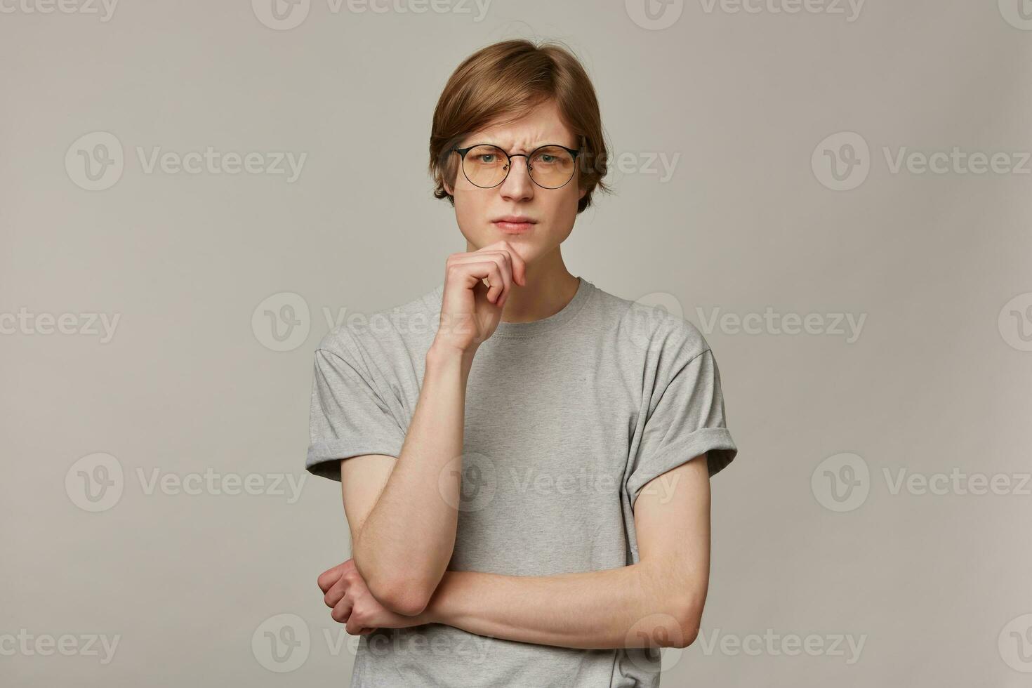 Portrait of serious, frowning male with blond hair. Wearing grey t-shirt and glasses. People and emotion concept. Touching his chin and thinks. Watching at the camera isolated over grey background photo
