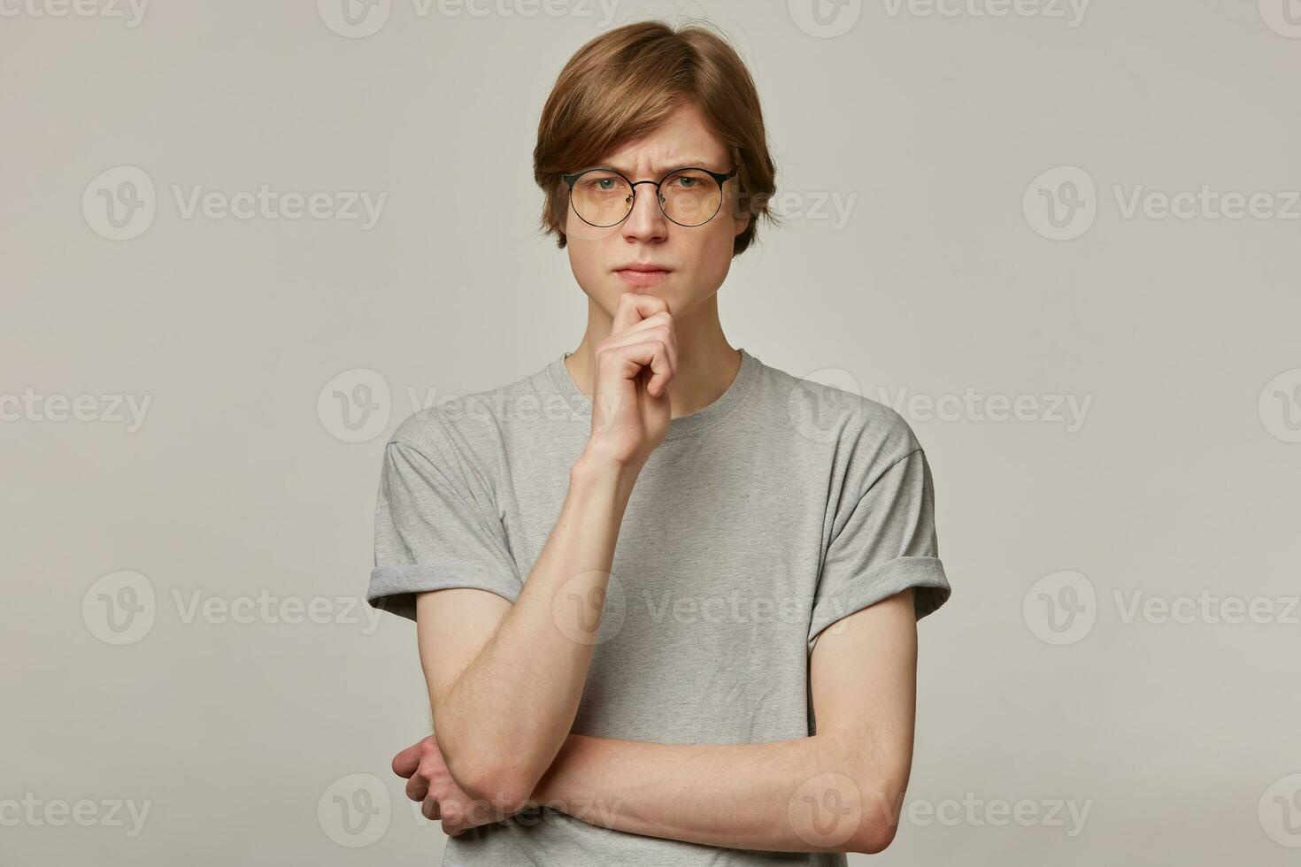 Serious looking male, thinking guy with blond hair. Wearing grey t-shirt and glasses. People and emotion concept. Frowns and touching his chin. Watching at the camera isolated over grey background photo