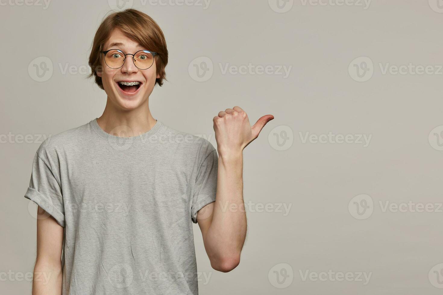 Teenage guy, happy looking man with blond hair. Wearing grey t-shirt, glasses and has braces. Watching at the camera and pointing with thumb to the right at copy space, isolated over grey background photo