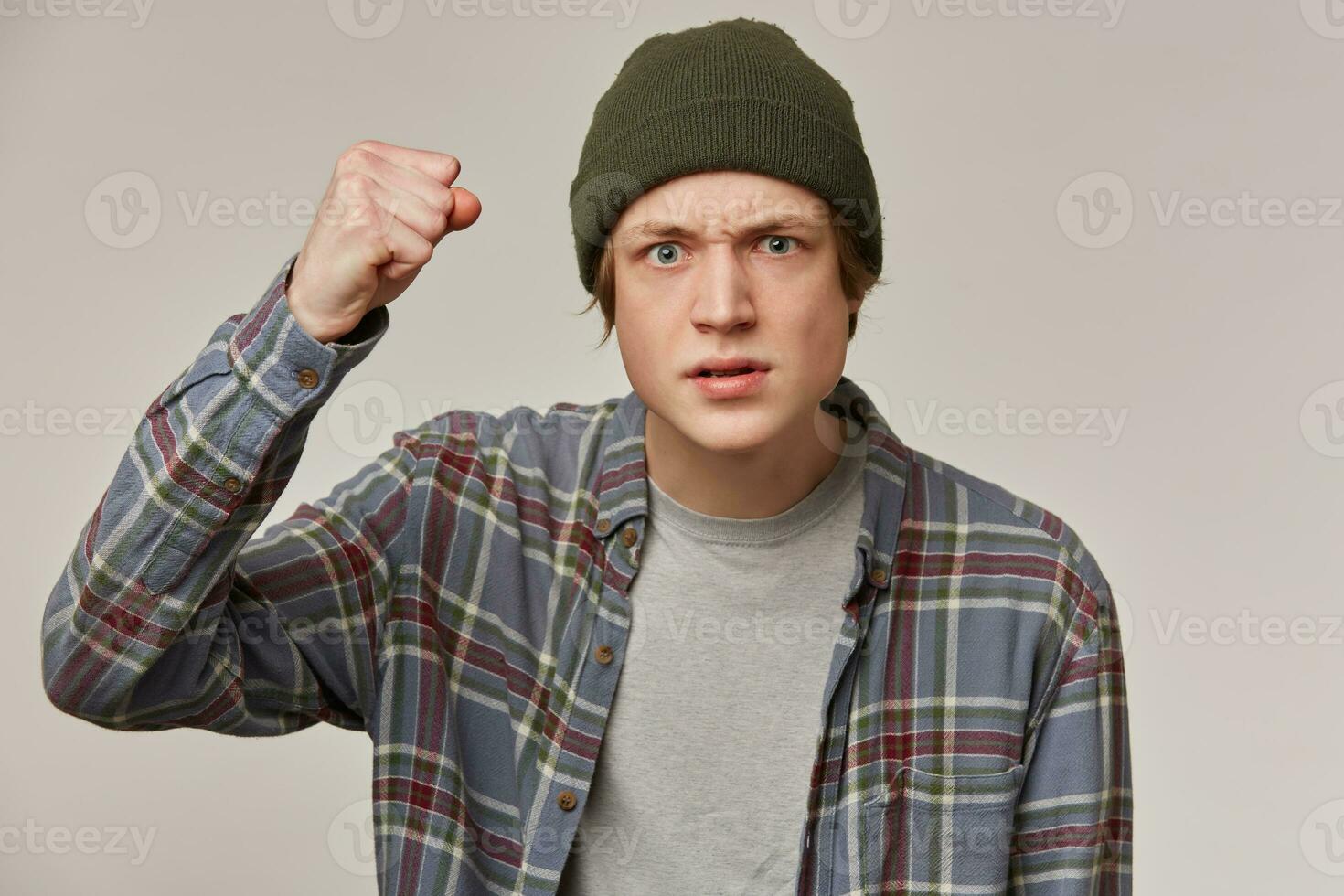 Frowning guy, angry looking man with blond hair. Wearing checkered shirt and beanie. People and emotion concept. Raises his fist, want to punch. Watching at the camera isolated over grey background photo