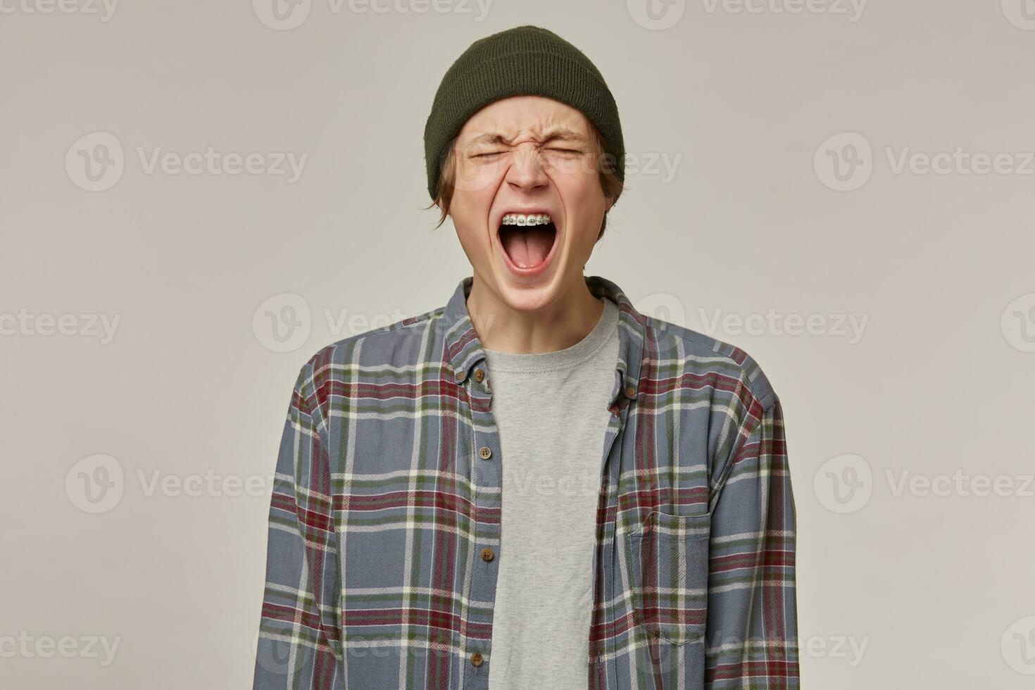 Teenage guy, unhappy looking man with blond hair. Wearing checkered shirt and beanie. Has braces. Keeps his eyes closed and yell. People and emotion concept. Stand isolated over grey background photo