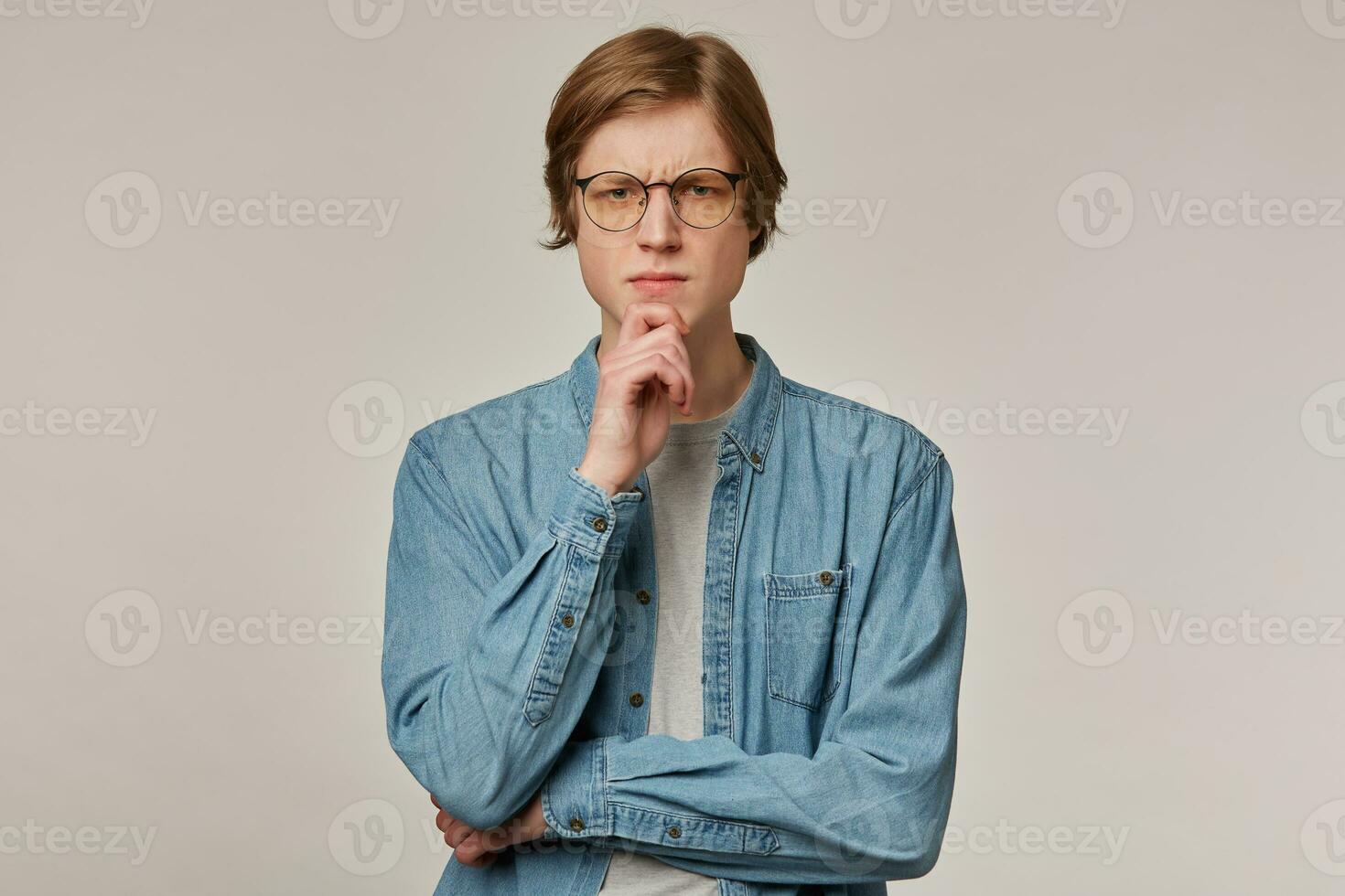 Cool looking male, focused guy with blond hair. Wearing denim shirt and glasses. Touching his chin and frowns. Emotion concept. Watching thoughtfully at the camera isolated over grey background photo