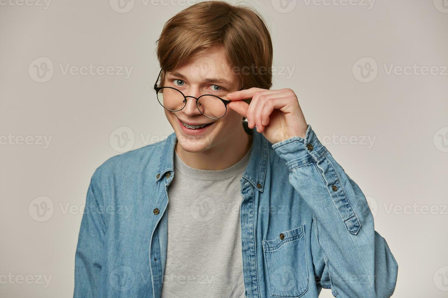 Cool looking male, handsome guy with blond hair. Wearing denim shirt and glasses. Has braces. Touching his eyewear and smiling. Emotion concept. Watching at the camera isolated over grey background photo