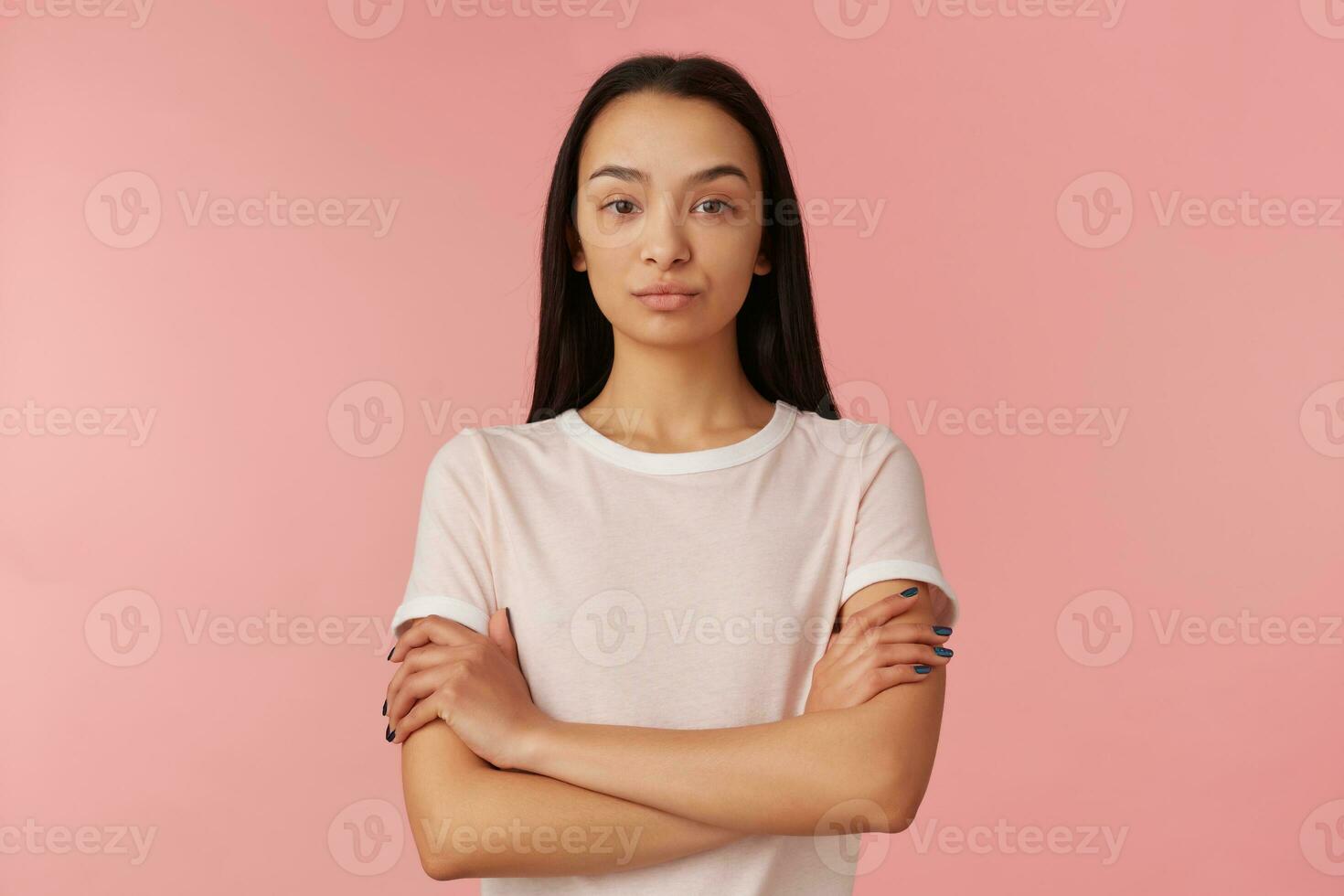 Portrait of serious, displeased girl with black long hair. Wearing white t-shirt. People and emotion concept. Keeps arms crossed, waiting. Watching at the camera isolated over pastel pink background photo