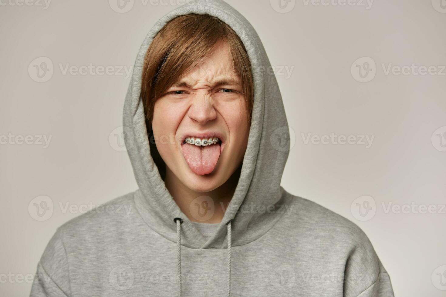 Portrait of displeased, adult male with blond hair. Wearing grey hoodie and has braces. Puts hood on and showing tongue. Emotion concept. Watching frowning at the camera isolated over grey background photo