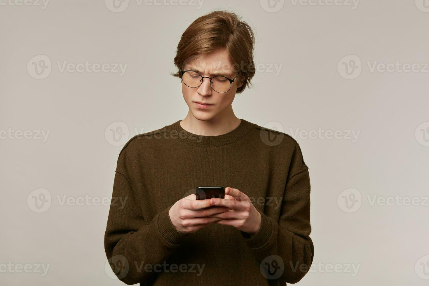 Serious looking male, handsome guy with blond hair. Wearing brown sweater and glasses. Holding a smartphone and frowns from what he read. Stand isolated over grey background photo