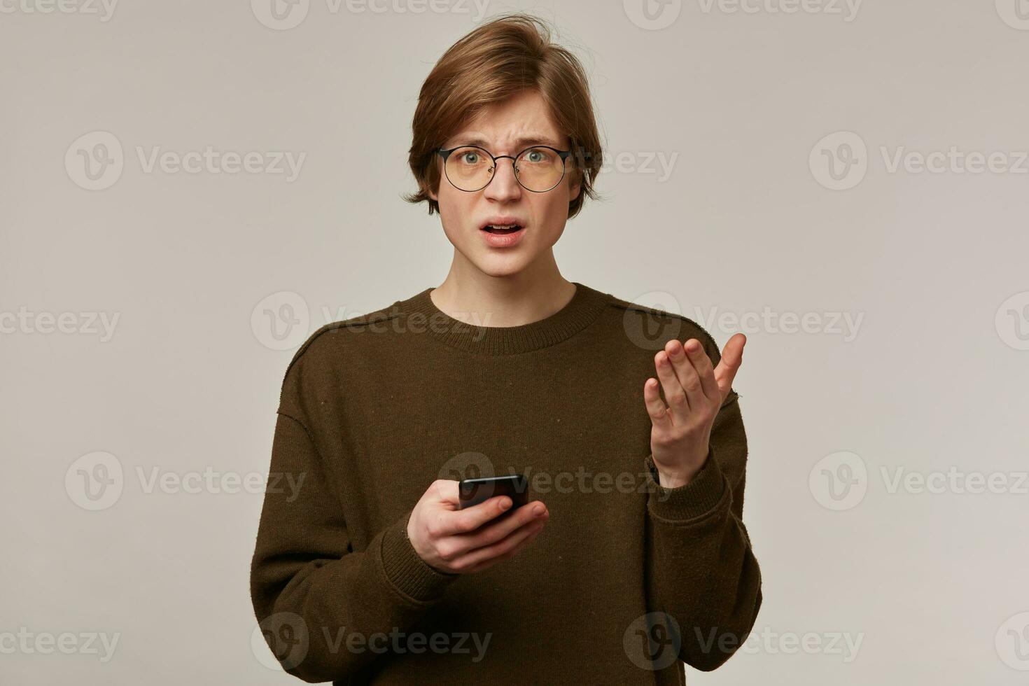Portrait of discontent, unhappy male with blond hair. Wearing brown sweater and glasses. Holding a smartphone. Confused from what he sees. Watching frowning at the camera isolated over grey background photo