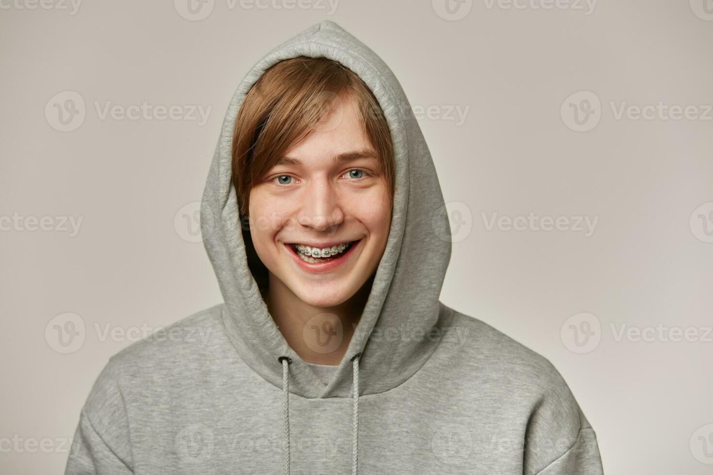 Cheerful teenage guy, happy looking man with blond hair. Wearing grey hoodie and has braces. Puts hood on and smiling. People and emotion concept. Watching at the camera isolated over grey background photo