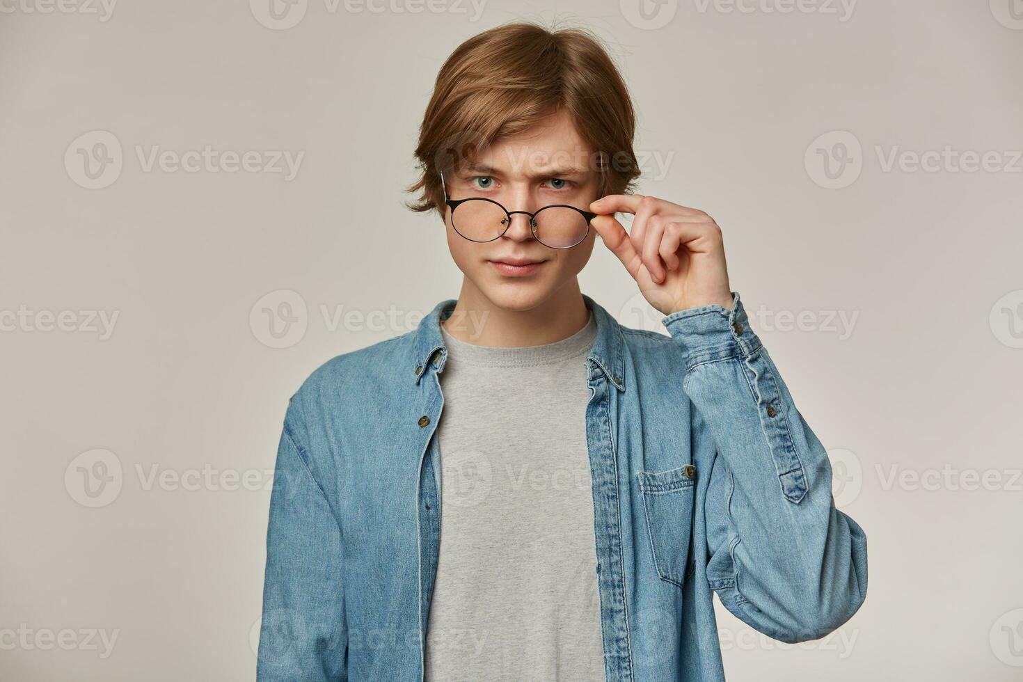 Portrait of serious, frowning male with blond hair. Wearing denim shirt and eyewear. Touching his glasses. Emotion concept. Watching accusingly at the camera isolated over grey background photo