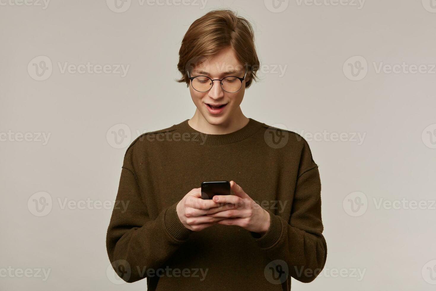 Teenage guy, happy looking man with blond hair. Wearing brown sweater and glasses. Holding a smartphone and looking at it. Typing a message. Stand isolated over grey background photo
