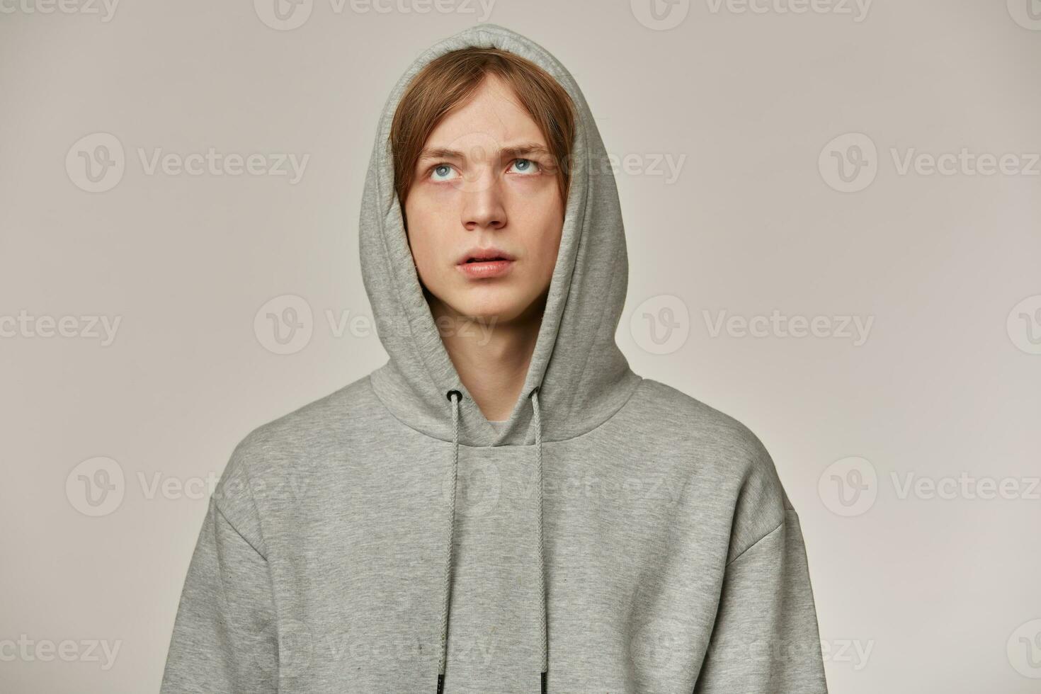 Displeased guy, bored looking man with blond hair. Wearing grey hoodie, puts hood on. Rolls his eyes. Annoyed about something. People and emotion concept. Stand isolated over grey background photo