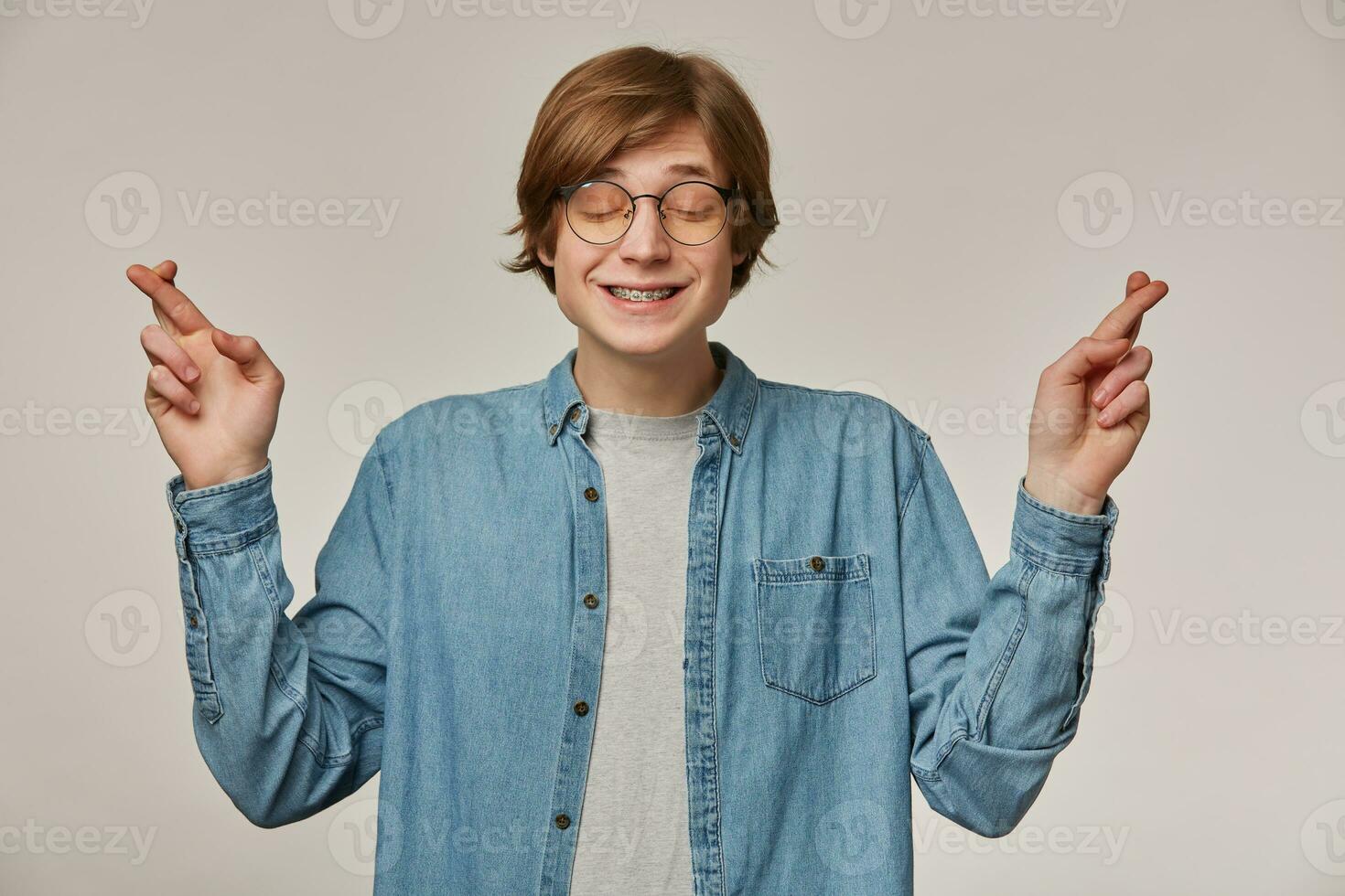 Teenage guy, happy looking man with blond hair. Wearing blue denim shirt, glasses and has braces. Cross his fingers and keeps eyes closed. Making a wish. Stand isolated over grey background photo