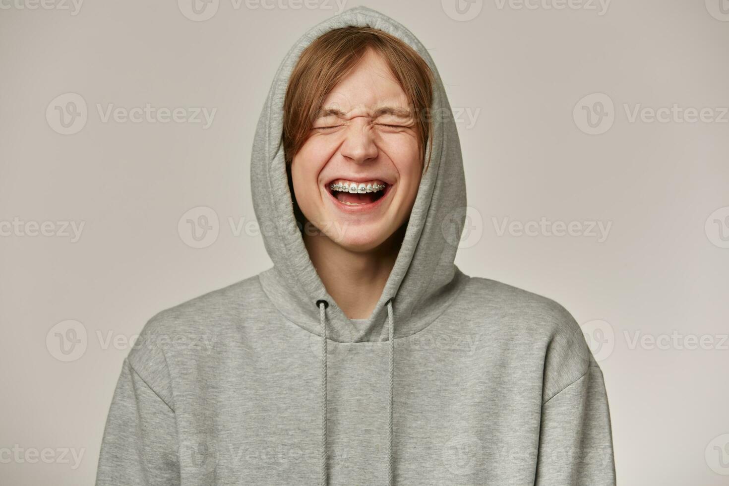 Cheerful male, handsome guy with blond hair. Wearing grey hoodie. Has braces. People and emotion concept. Puts hood on and laughing with closed eyes. Stand isolated over grey background photo