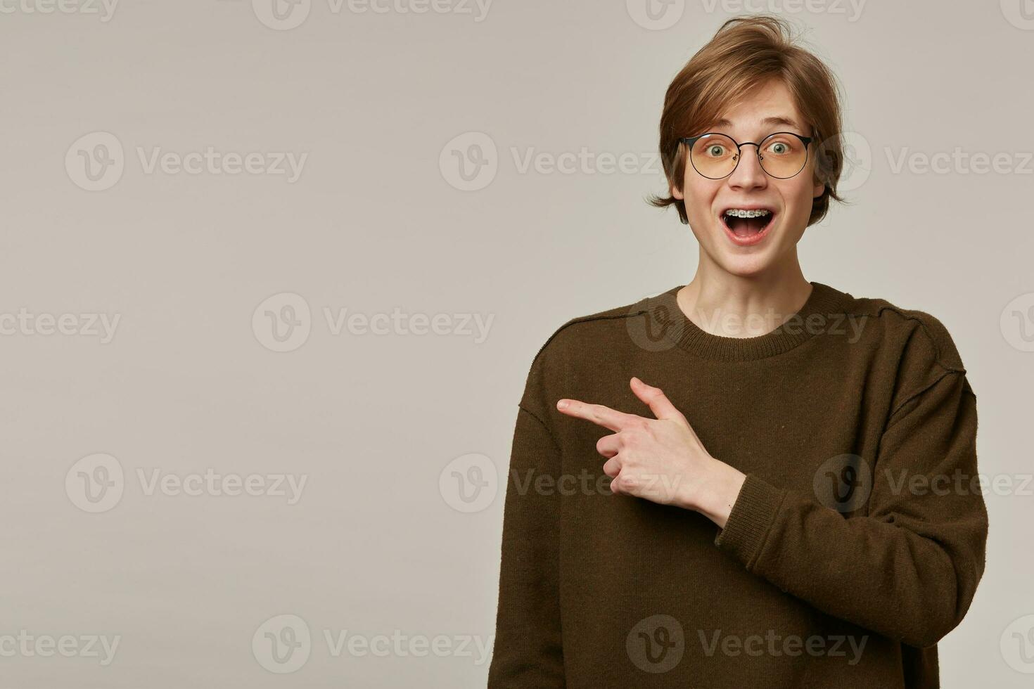 Teenage guy, amazed looking man with blond hair. Wearing brown sweater, glasses and has braces. Watching at the camera and pointing finger to the left at copy space, isolated over grey background photo