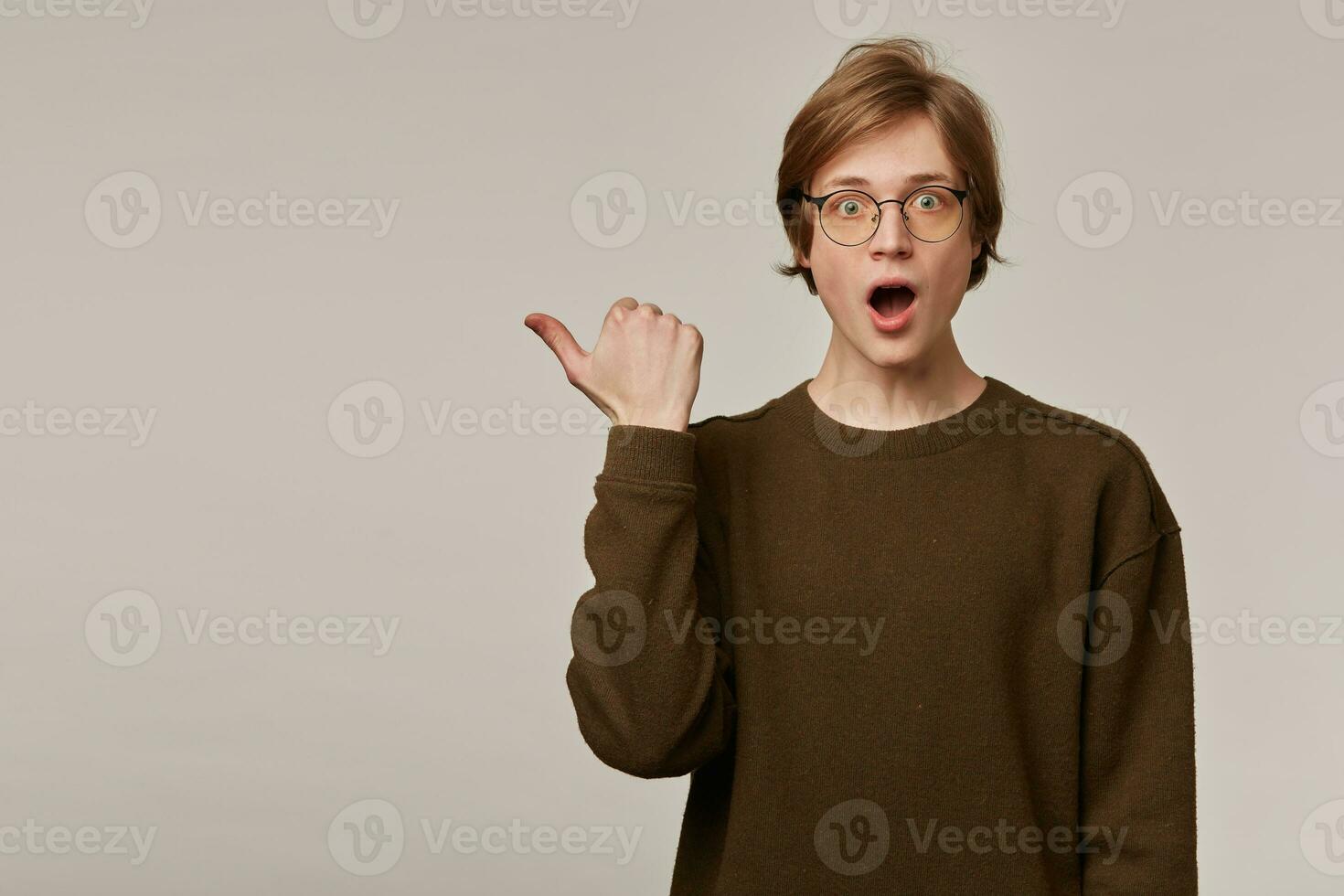 Shocked looking male, handsome guy with blond hair. Wearing brown sweater and glasses. Watching at the camera and pointing thumb to the left at copy space, isolated over grey background photo