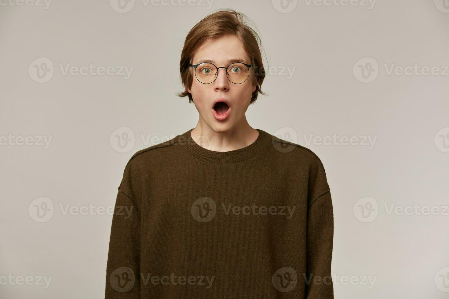 Shocked looking male, handsome guy with blond hair. Wearing brown sweater and glasses. Opens mouth in surprise. People and emotion concept. Watching at the camera isolated over grey background photo