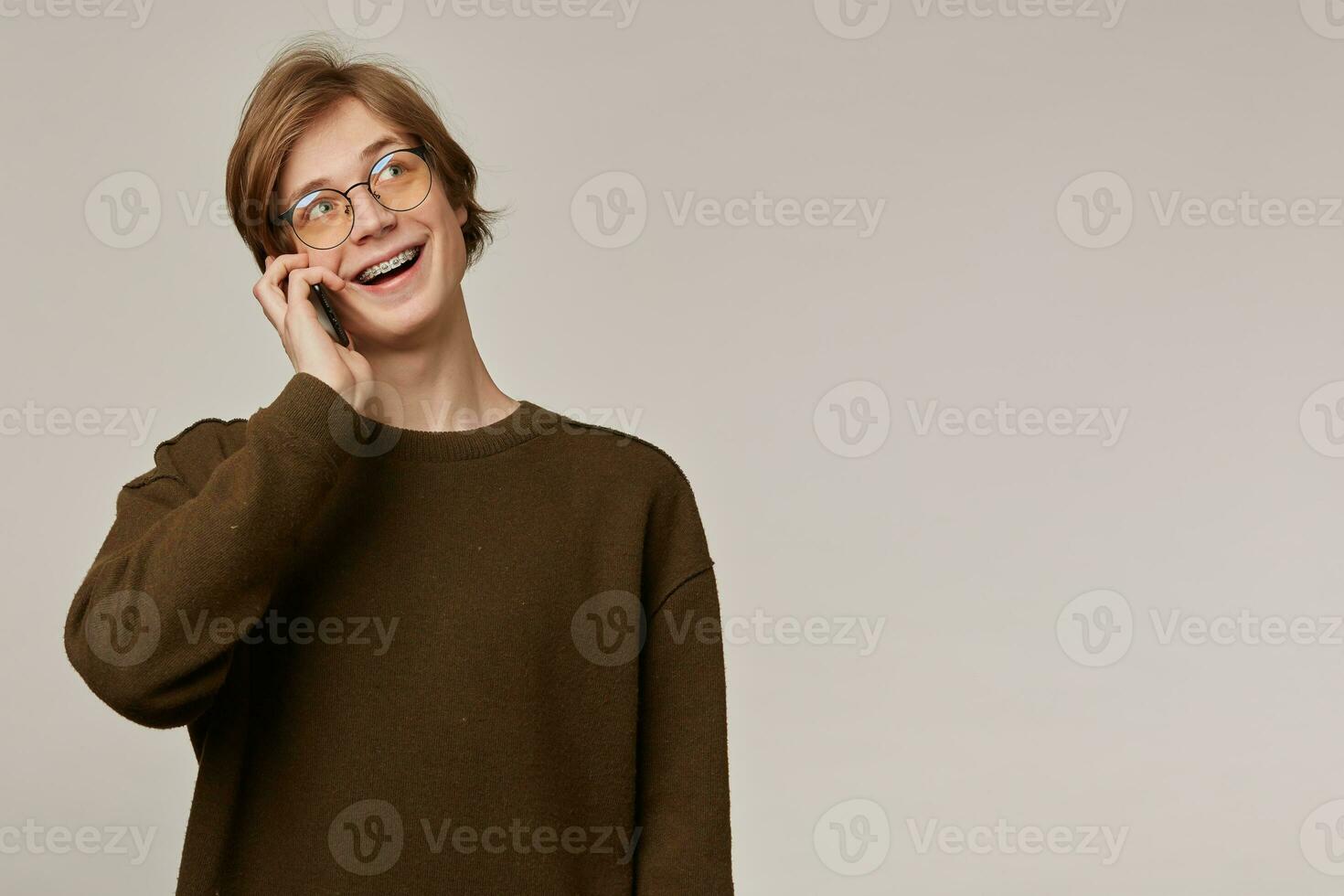 Cheerful male, handsome guy with blond hair. Wearing brown sweater and glasses. Has braces. Talk on a phone, pleasant conversation. Watching to the right at copy space, isolated over grey background photo