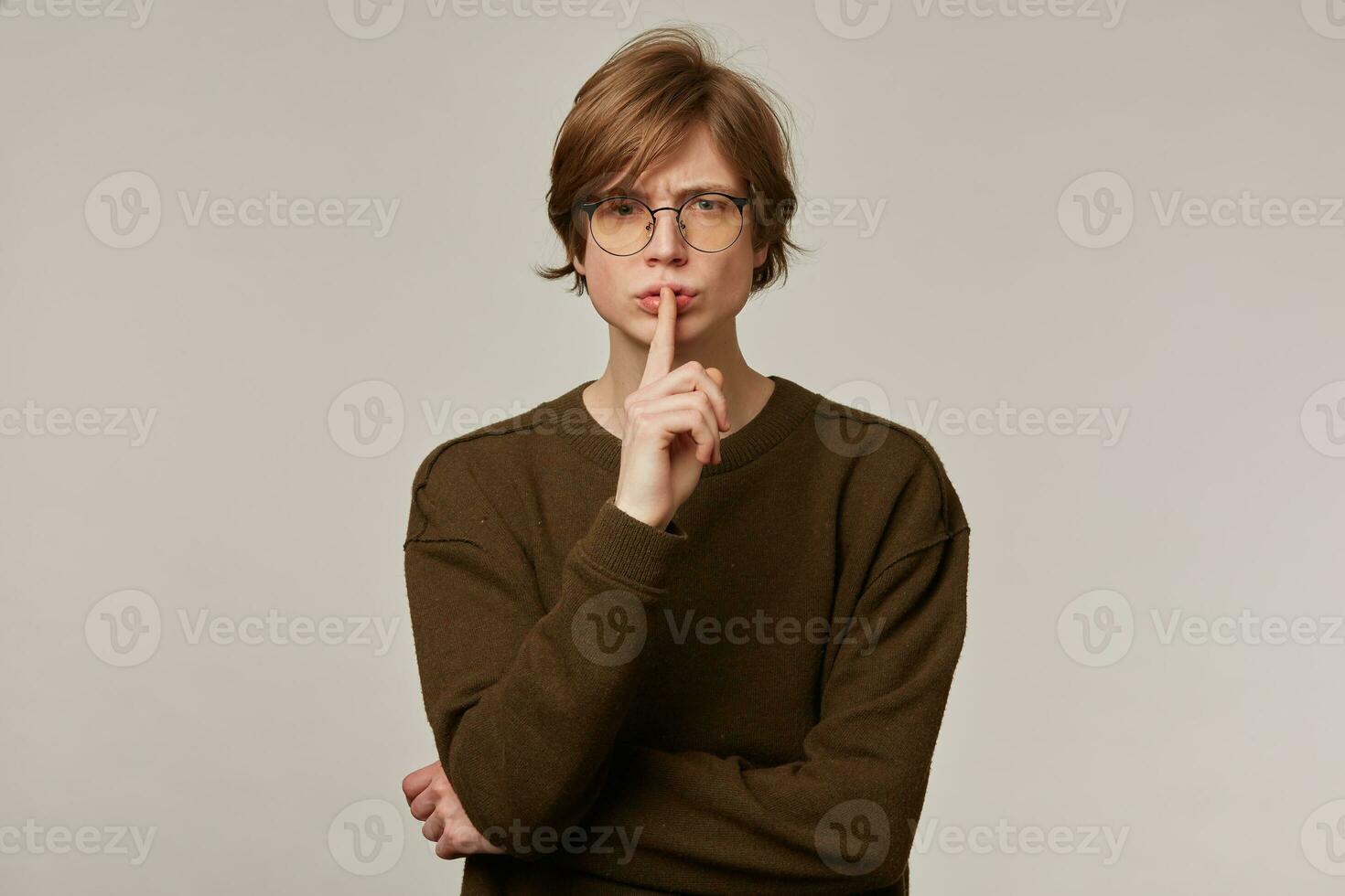 Portrait of handsome, serious male with blond hair. Wearing brown sweater and glasses. Showing silence sign. People and emotion concept. Watching at the camera isolated over grey background photo