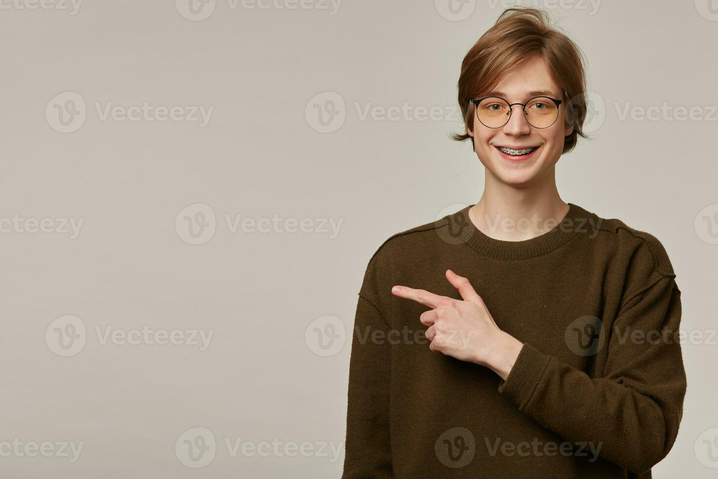 Portrait of positive, adult male with blond hair. Wearing brown sweater and glasses. Has braces. Watching at the camera and pointing finger to the left at copy space, isolated over grey background photo