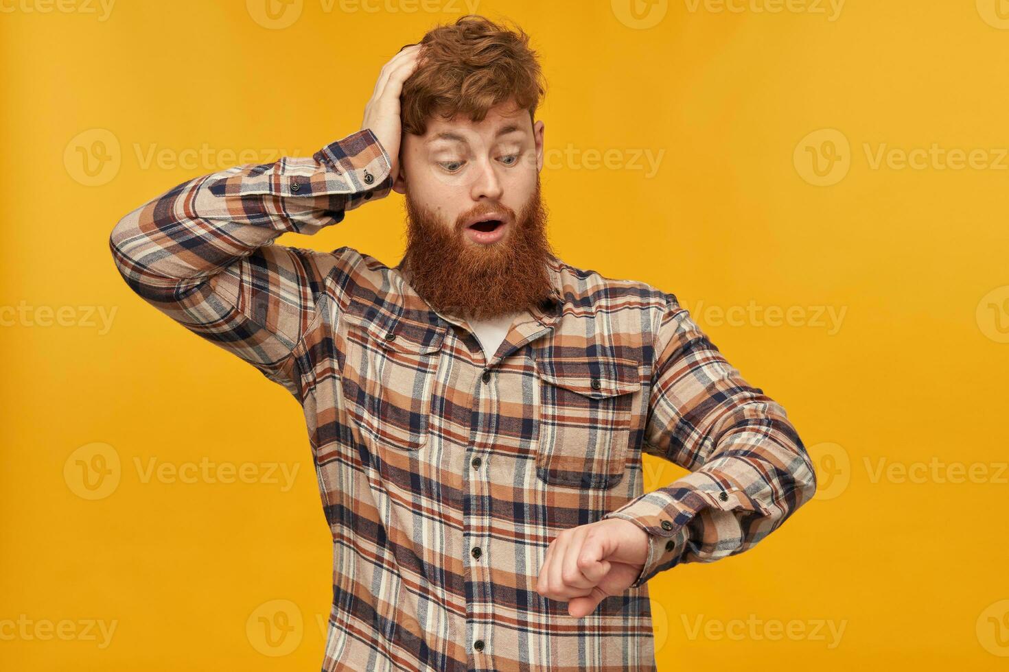 Young male student, with red hair and big beard, looks at his watches, keeps his hand on head and think about that he is late at work. Isolated over yellow background, photo