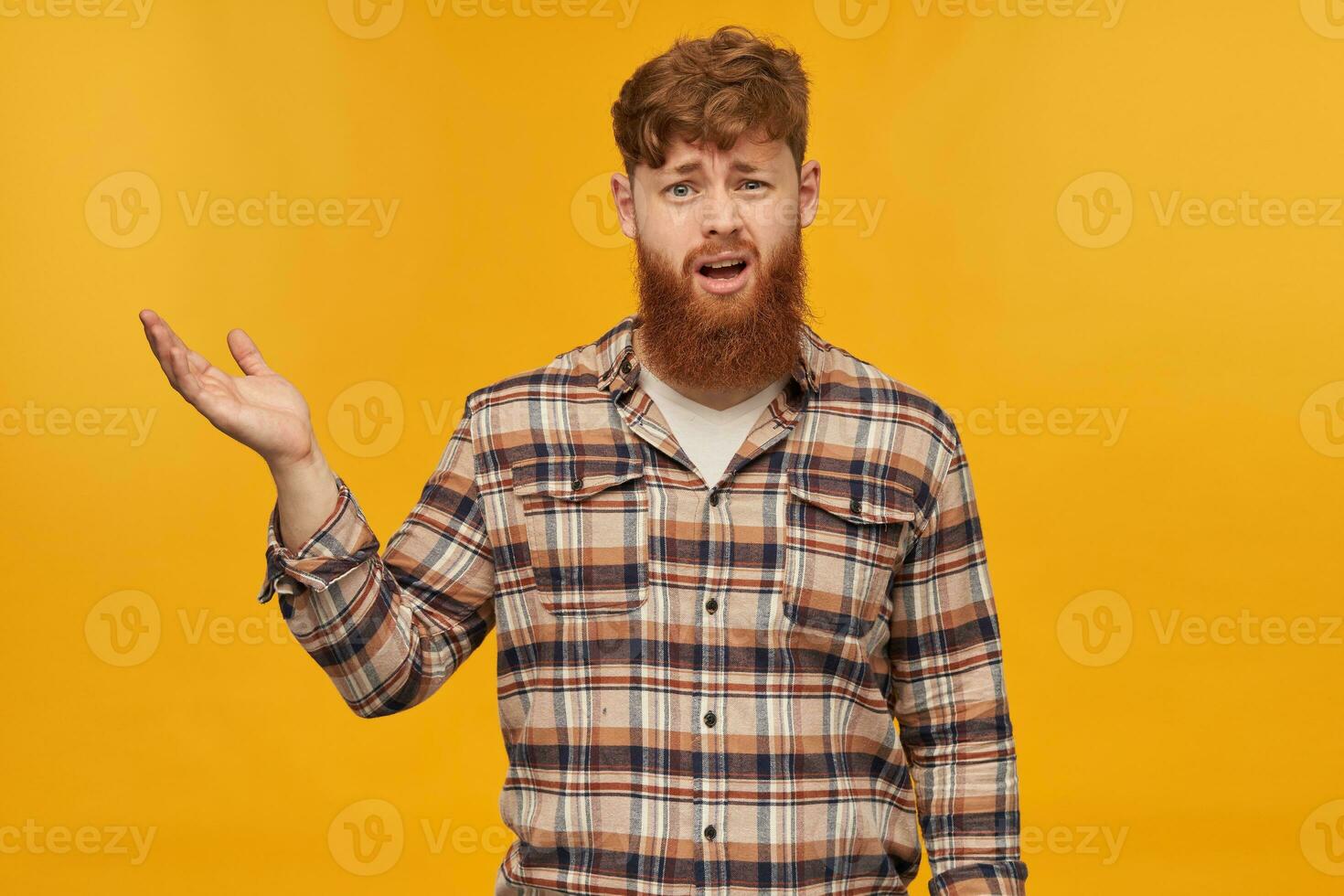 Indoor shot of sad depressed young male, looks into camera with confused facial expression, raised his hand, isolated over yellow background photo