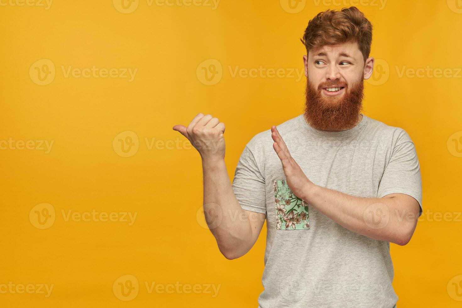 Indoor shot of unsure young male, wears grey t-shirt, with big beard and red hair, point with a finger left at copy space with negative facial expression. Shows stop sign. isolated photo